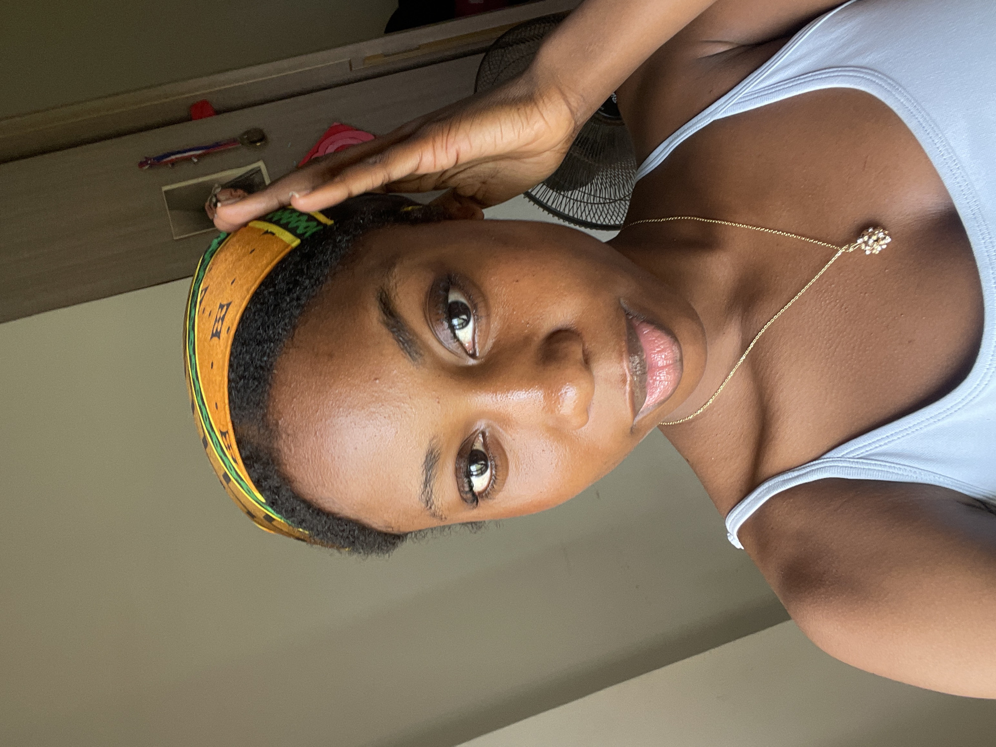 A close-up, slightly tilted portrait captures a person with dark skin and dark hair looking directly into the camera. They wear a light blue or white tank top and a delicate gold-colored necklace with a small, sparkling, flower-like pendant. A wide, colorful headband, featuring an orange-yellow base with green trim and a repeating dark brown, possibly stylized letter pattern, adorns their head. Their right hand is raised, with fingers touching their forehead, partly obscuring the top of their head. The person's expression is neutral, and their lips appear slightly glossy.
The background suggests an indoor setting, with soft, natural light illuminating the scene, particularly highlighting the person's skin. Visible in the blurred background are parts of a dark-colored fan and what appears to be a light-colored wall or piece of furniture. In the upper left corner, small, indistinct items are visible on a surface, including a red object and a metallic, key-like item. The overall impression is a casual, posed portrait taken indoors during the daytime.