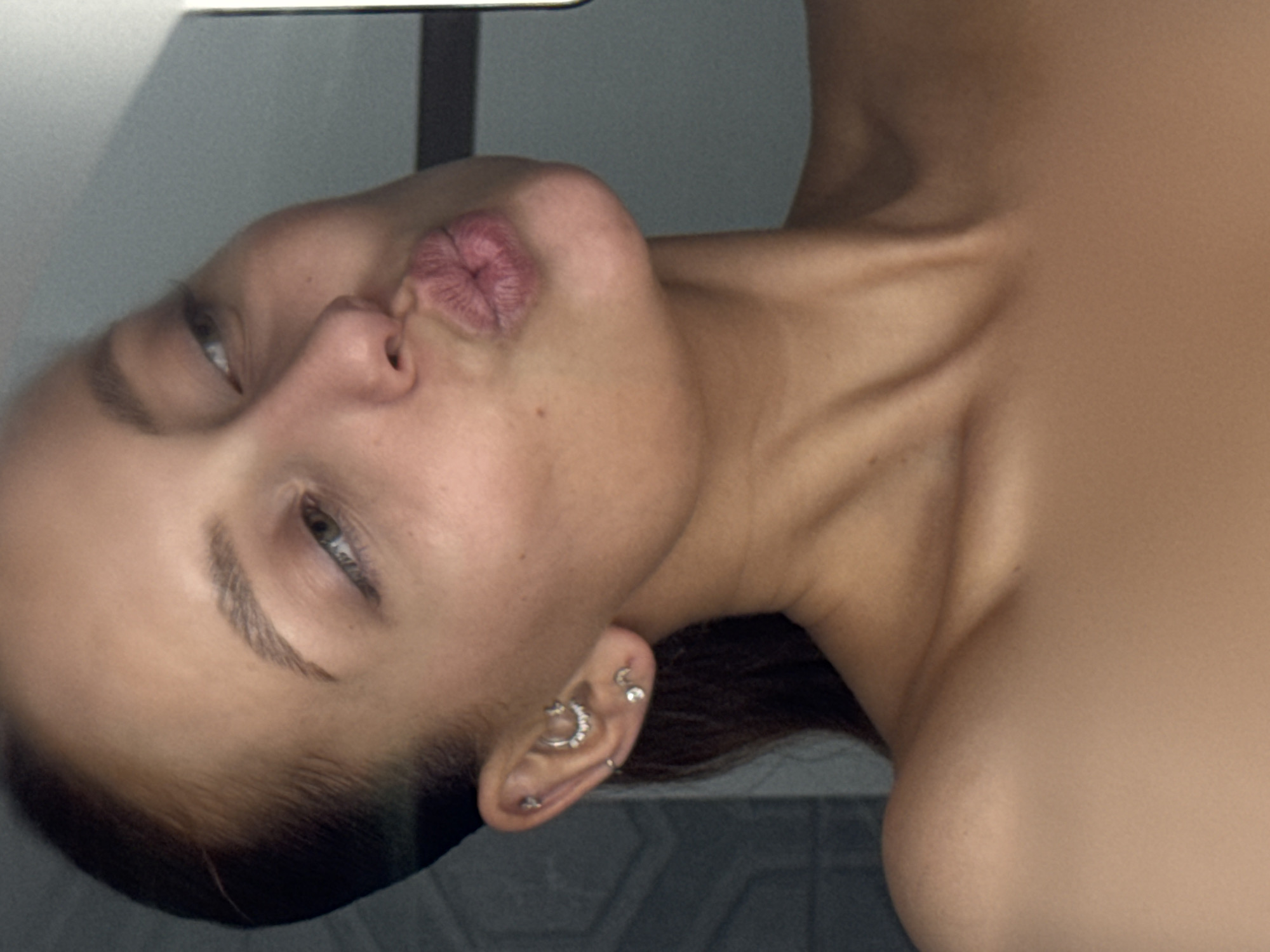 A close-up portrait shows a light-skinned person from the neck up, oriented with their head tilted significantly to the left. The person has pursed lips, fair skin, and light-colored eyes with visible eyelashes and eyebrows. The left ear, in view, has multiple piercings, including a larger ring and several smaller studs. The background appears to be a neutral gray, possibly a wall or tile. The person's shoulder and neck are visible on the right side of the frame. The image is well-lit and relatively sharp. The location, Chaikovsky, Russia, is not directly identifiable in the image.
