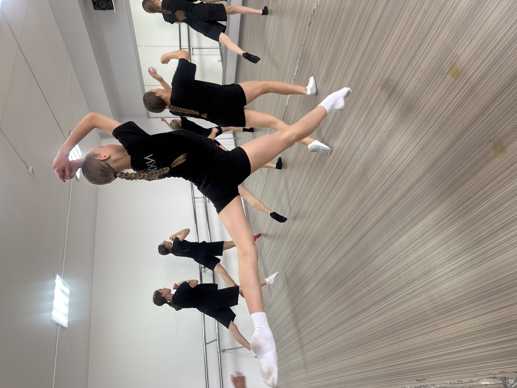 This media file shows a group of young women in a dance studio in Votkinsk, Russia. They are all wearing black leotards and shorts, and some are wearing white socks. The dance studio has a wooden floor and a ballet barre along one wall. The lighting is bright, suggesting it's daytime. The women appear to be in the middle of a dance class or rehearsal, as they are in various poses, some with arms extended and legs lifted. One woman in the foreground is captured mid-movement, with her leg extended high and her arms gracefully positioned. The overall mood appears to be one of focused activity and athleticism. There is text visible on the leotard of one of the dancers, but it is partially obscured and unreadable. The scene suggests a structured environment dedicated to dance training.
