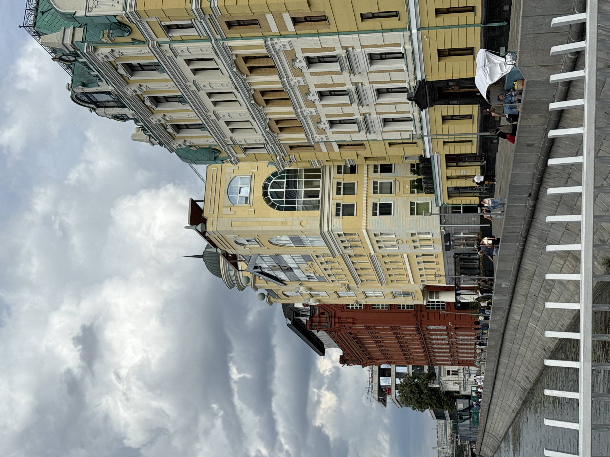 The image shows a row of buildings along a river. The buildings are of various architectural styles and colors, with the most prominent ones being red brick and yellow stucco. There is a promenade or walkway in front of the buildings, with people walking and sitting. A white umbrella shelter is visible on the promenade. The river is calm, and the sky is cloudy. The scene suggests an urban waterfront environment.