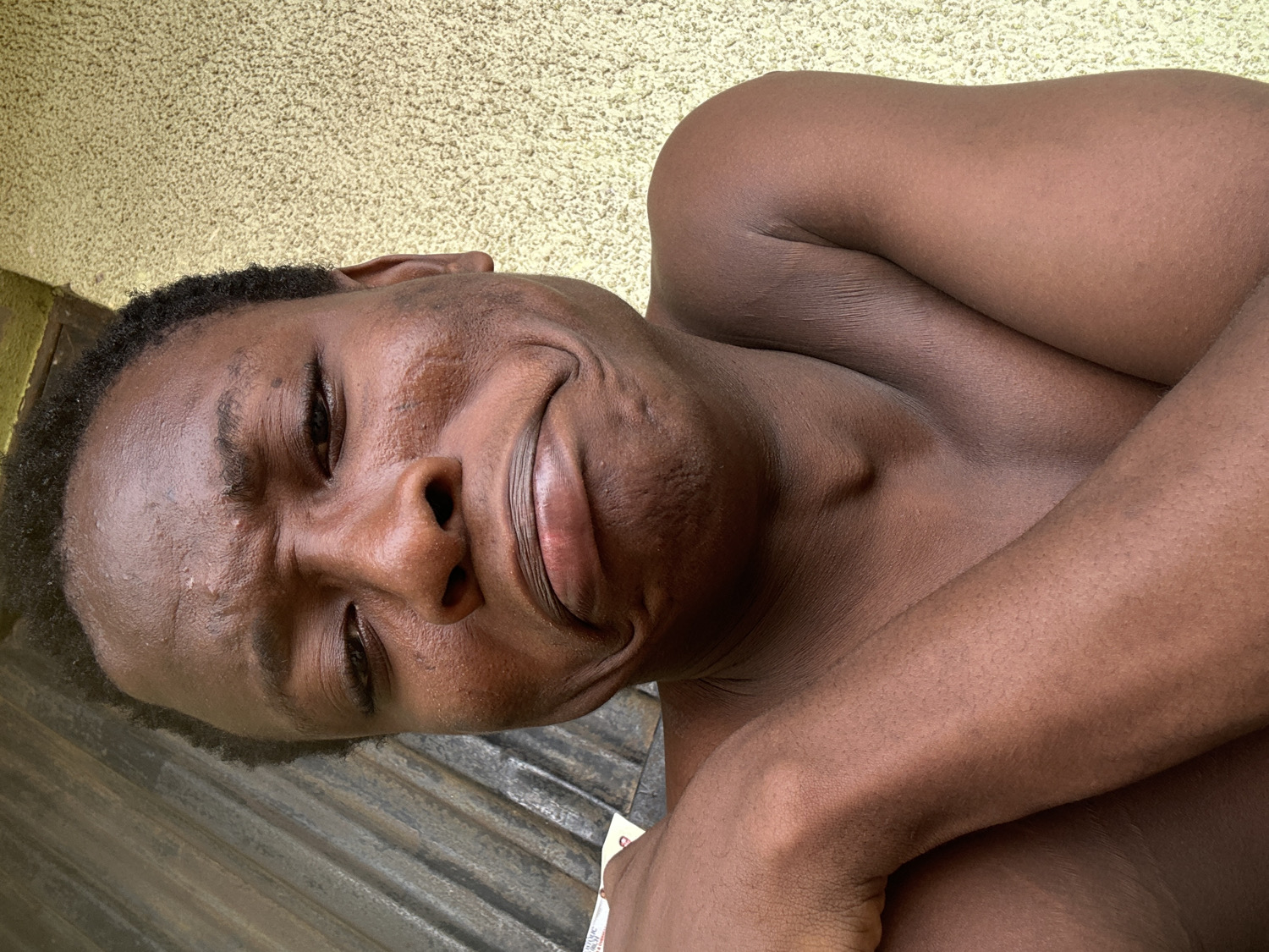 A young Black man is depicted in a close-up shot, appearing shirtless. His head is positioned towards the left side of the frame, tilted slightly upwards, with his body extending to the right. He has short, dark, curly hair and dark skin, which appears somewhat dewy or shiny.
He is captured making a playful and expressive face: his eyes are partially closed, eyebrows furrowed, and his mouth is pursed in an exaggerated pout or smirk. His left arm is visible, bent at the elbow, and part of his bare chest is also in view.
The background consists of two distinct textures: a light yellow or beige textured wall (possibly stucco or rough plaster) is seen above his head, while a darker, corrugated or grooved surface, resembling metal or weathered wood paneling, is visible below him. The lighting suggests it is daytime. In the bottom right corner, a small portion of a white object, possibly paper, is visible with faint, illegible markings on it. The overall mood conveyed by the man's expression is humorous and engaging.