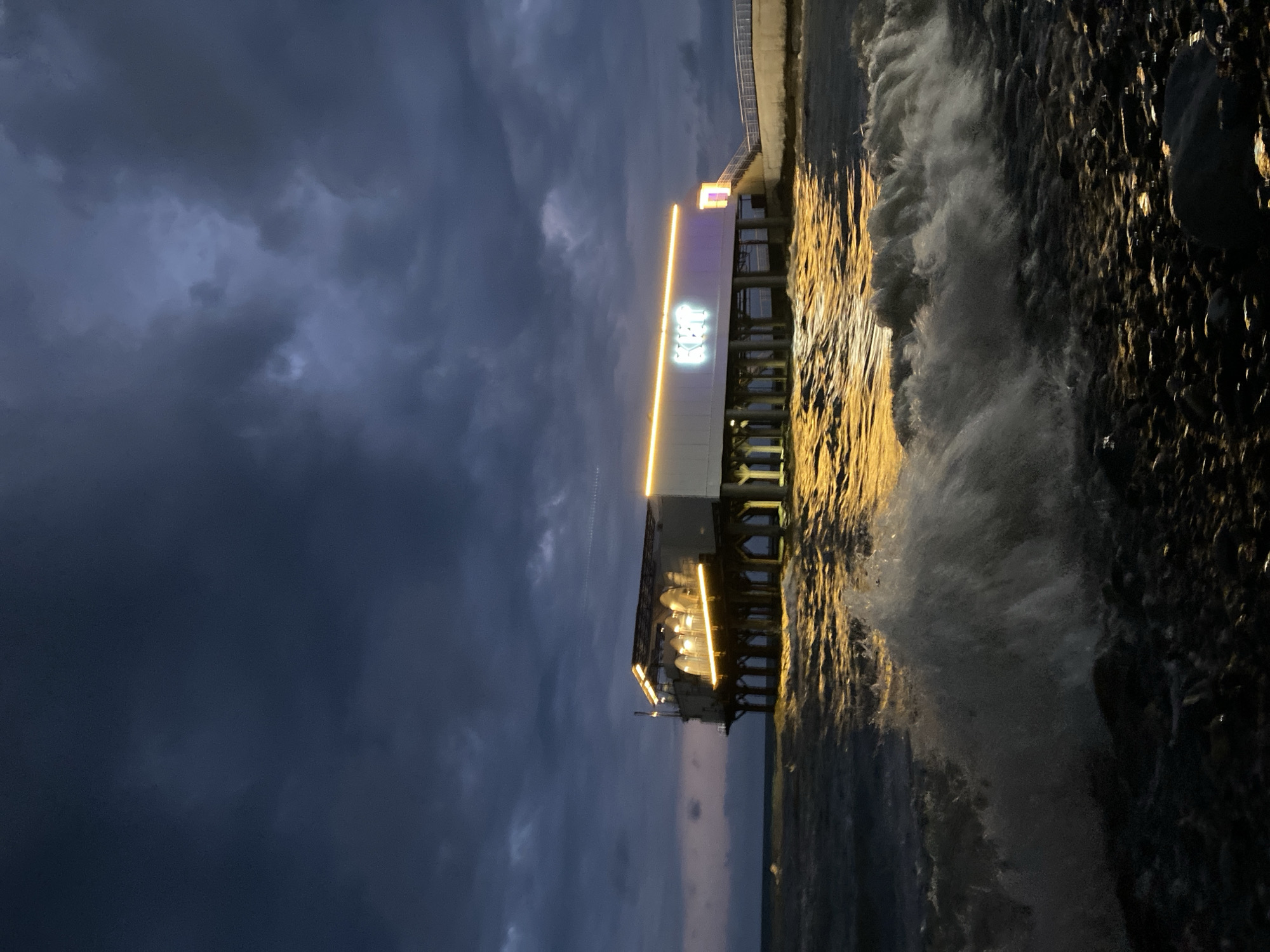 The image shows a pier at night, likely in Sochi, Russia. The structure is illuminated by golden lights that reflect on the churning water. There is a building on the pier with the word "Sochi" written in Cyrillic, further suggesting the location. The sky is a dark, cloudy blue, creating a dramatic contrast with the brightly lit pier and water. Waves crash against the pier supports, adding to the dynamic energy of the scene.