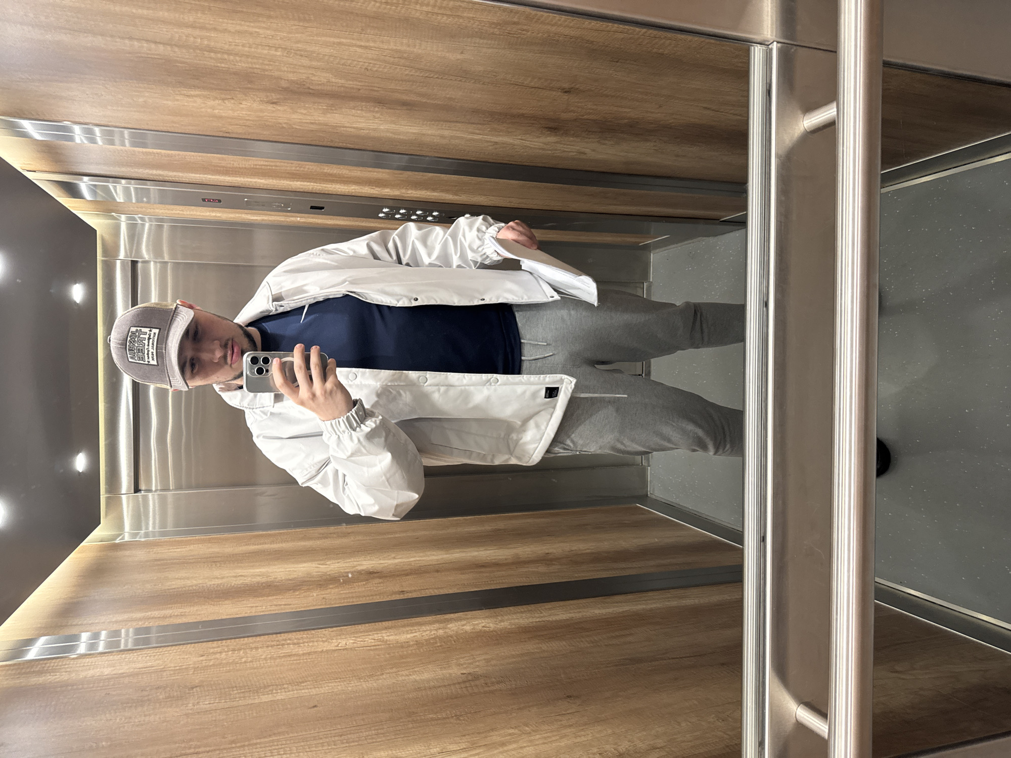 A young man takes a mirror selfie in an elevator, holding a silver smartphone with his right hand. He is wearing a light-colored baseball cap with a rectangular patch displaying mirrored text, which appears to say "THE ORIGINAL 2007". He is dressed in a white track jacket, a dark navy t-shirt, and grey sweatpants. His expression is neutral.
The elevator interior features polished wooden panels on the side walls, accented by horizontal silver metal strips. The back wall is made of brushed stainless steel. A control panel with multiple illuminated white buttons, indicating floor selections, is visible on the stainless steel wall. Stainless steel handrails are also present on the right side of the elevator, extending vertically. The floor of the elevator is dark grey, possibly with a subtle speckled texture. The lighting inside the elevator appears to be artificial, typical of an indoor setting. The image is taken in Sofia, Bulgaria.