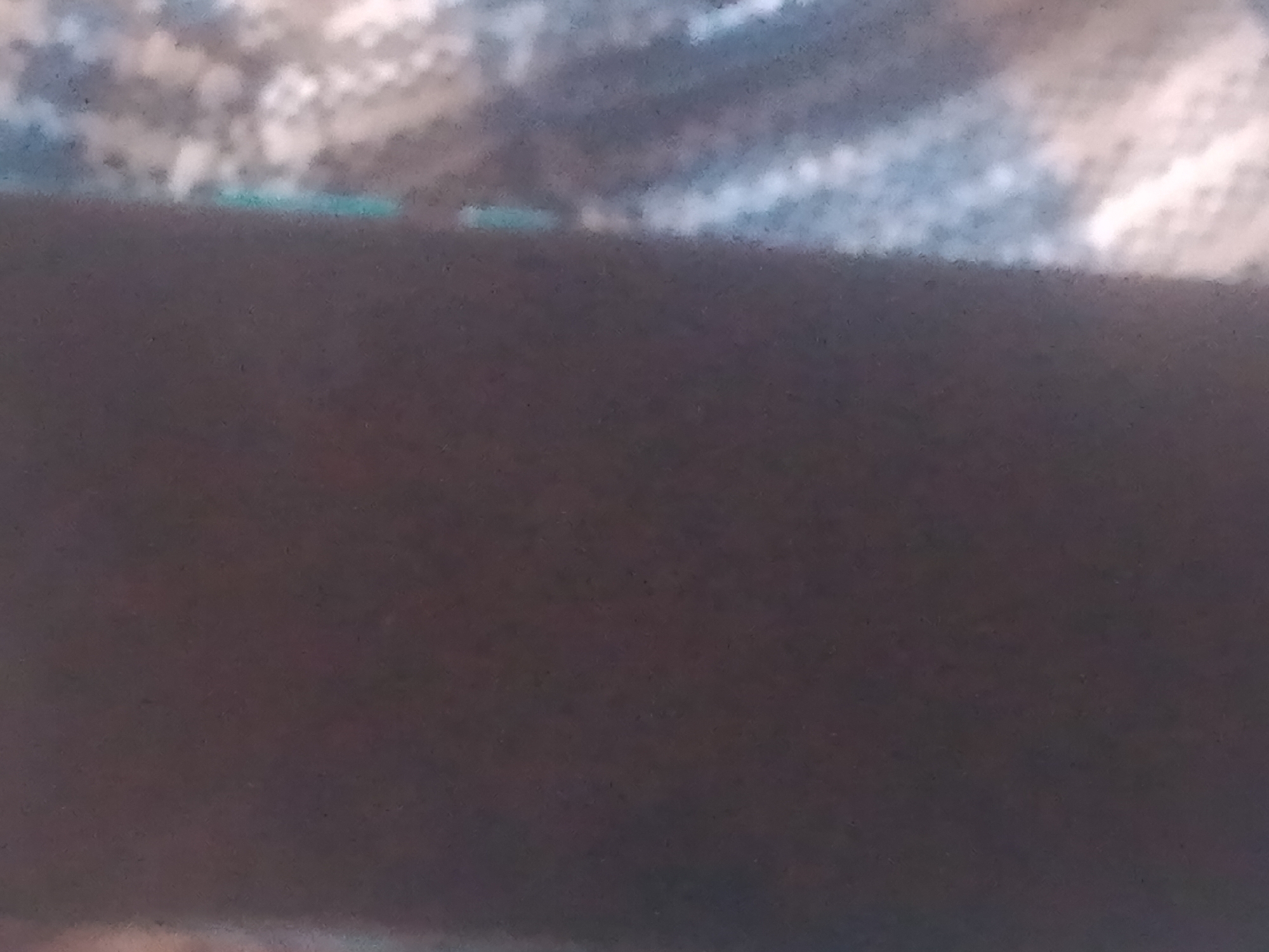 The image shows a close-up, out-of-focus view of a dark, textured surface that appears to be a piece of fabric. Above this dark surface, there is a blurry band of lighter colors, suggesting a different material or pattern. The lighting is soft and diffused, making it difficult to discern specific details about the objects or their surroundings. There are no discernible people, actions, or interactions visible in this image, and the context of Jauro Yinu, Nigeria is not evident from the visual information provided.