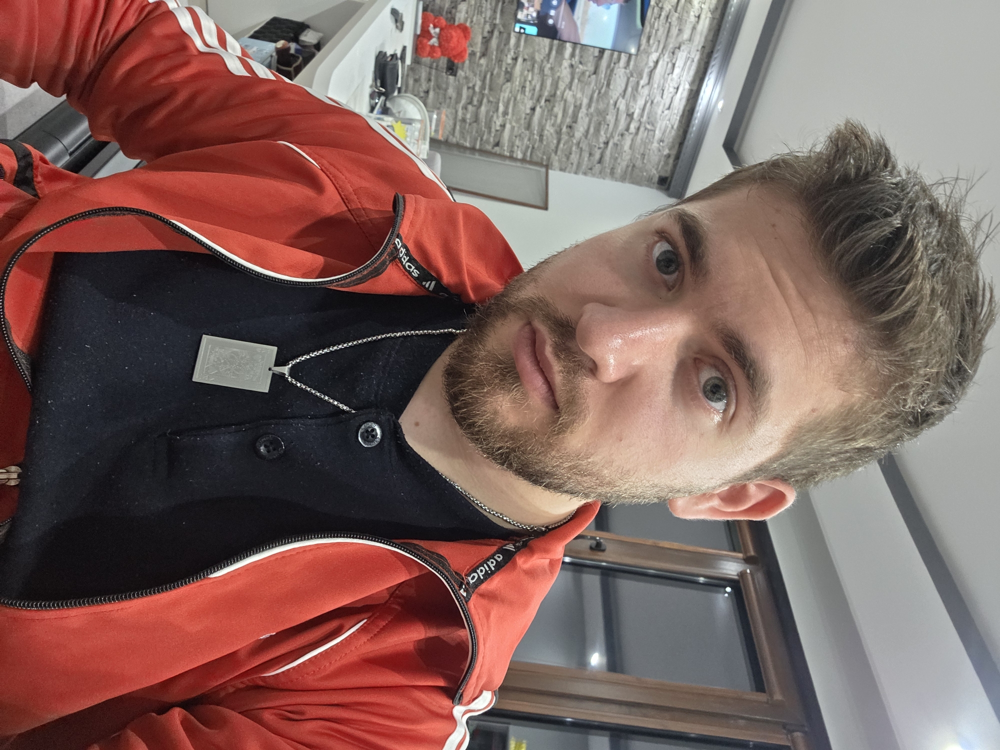 A close-up, slightly angled, indoor photograph depicts a man with light brown hair and a beard. He wears a red Adidas tracksuit jacket over a dark-colored shirt with several buttons. A silver chain with a rectangular pendant hangs around his neck. The background includes a light gray wall, a television screen, and a window. There is no information to suggest the location is Yagodovo, Bulgaria.