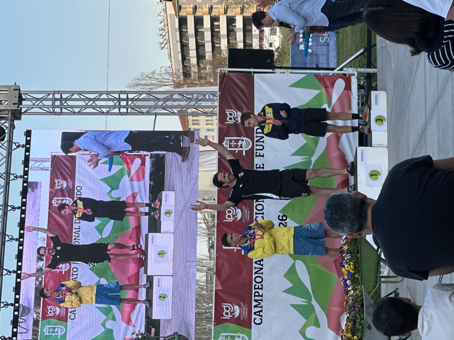 This image depicts an outdoor awards ceremony taking place in Mirandela, Portugal. On a central stage, three male athletes are positioned on a three-tiered podium, numbered "1" (center), "2" (left), and "3" (right), with "MIRANDELA" visible on the podium steps. A large LED screen directly above the stage mirrors the live scene below.
The athlete on the first-place step (center) is a man with dark hair and a beard, dressed in a black short-sleeved top, black shorts, and black sandals. He has both arms raised in a celebratory gesture and is smiling, wearing a medal around his neck. The athlete on the second-place step (viewer's left) is a young man in a yellow long-sleeved jacket, blue shorts, and light-colored athletic shoes. He holds a medal in front of him. The athlete on the third-place step (viewer's right) is a young man in a dark blue and yellow long-sleeved top, black shorts, and orange and grey athletic shoes, also holding a medal.
To the right of the podium, an official, a man in a light blue collared shirt and dark trousers, stands clapping, with a blue object in his left hand. A large banner behind the podium displays the text "CAMPEONATO NACIONAL DE FUNDO," along with the logos of IPDJ and FPC, and stylized green and white mountain graphics. A black speaker is visible to the right of the stage. To the left of the podium, a green wheelbarrow holds a collection of colorful potted plants.
In the immediate foreground, the back of a man's head and shoulders, wearing a black t-shirt, is visible, along with parts of other spectators to his left and right. The background features multi-story urban buildings and some bare trees under a clear, bright blue sky, indicating daylight conditions.