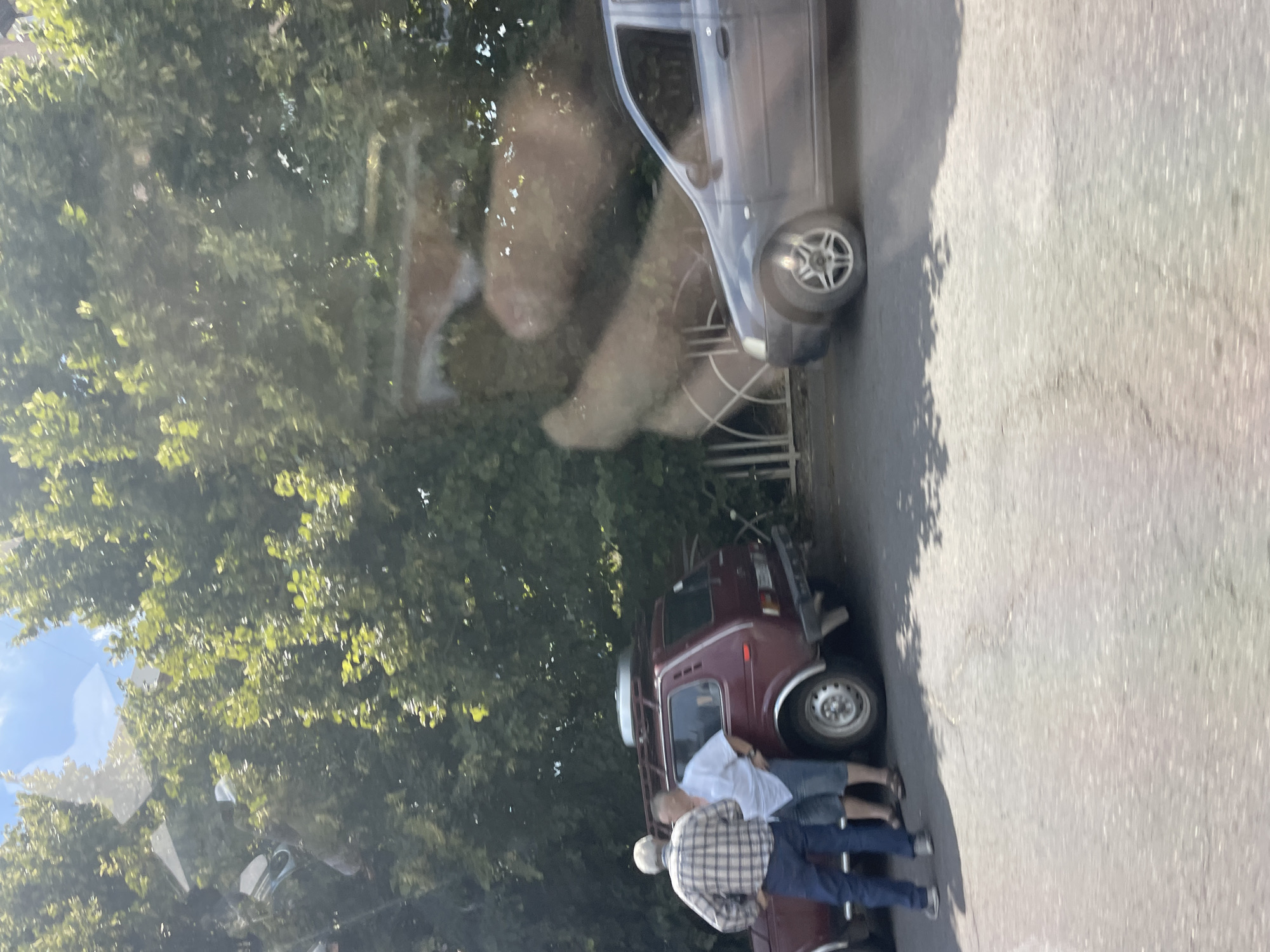 The image depicts a street scene with two men standing next to a maroon-colored Lada Niva vehicle. One man, wearing a plaid shirt and blue jeans, is facing away from the camera. The other man, in a white t-shirt and shorts, is facing the first man and appears to be engaged in conversation. The Lada Niva has a roof rack with a silver object on top and is parked on an asphalt surface. To the right of the Lada Niva, a dark gray sedan is visible, also parked on the asphalt. Behind the vehicles, there is dense green foliage from trees, suggesting a location with ample vegetation. The sky above is partially visible with some clouds and sunlight filtering through the leaves, indicating daytime. The overall environment appears to be an outdoor urban or semi-urban setting. The image seems to be taken from within a vehicle, evidenced by reflections on the surfaces.