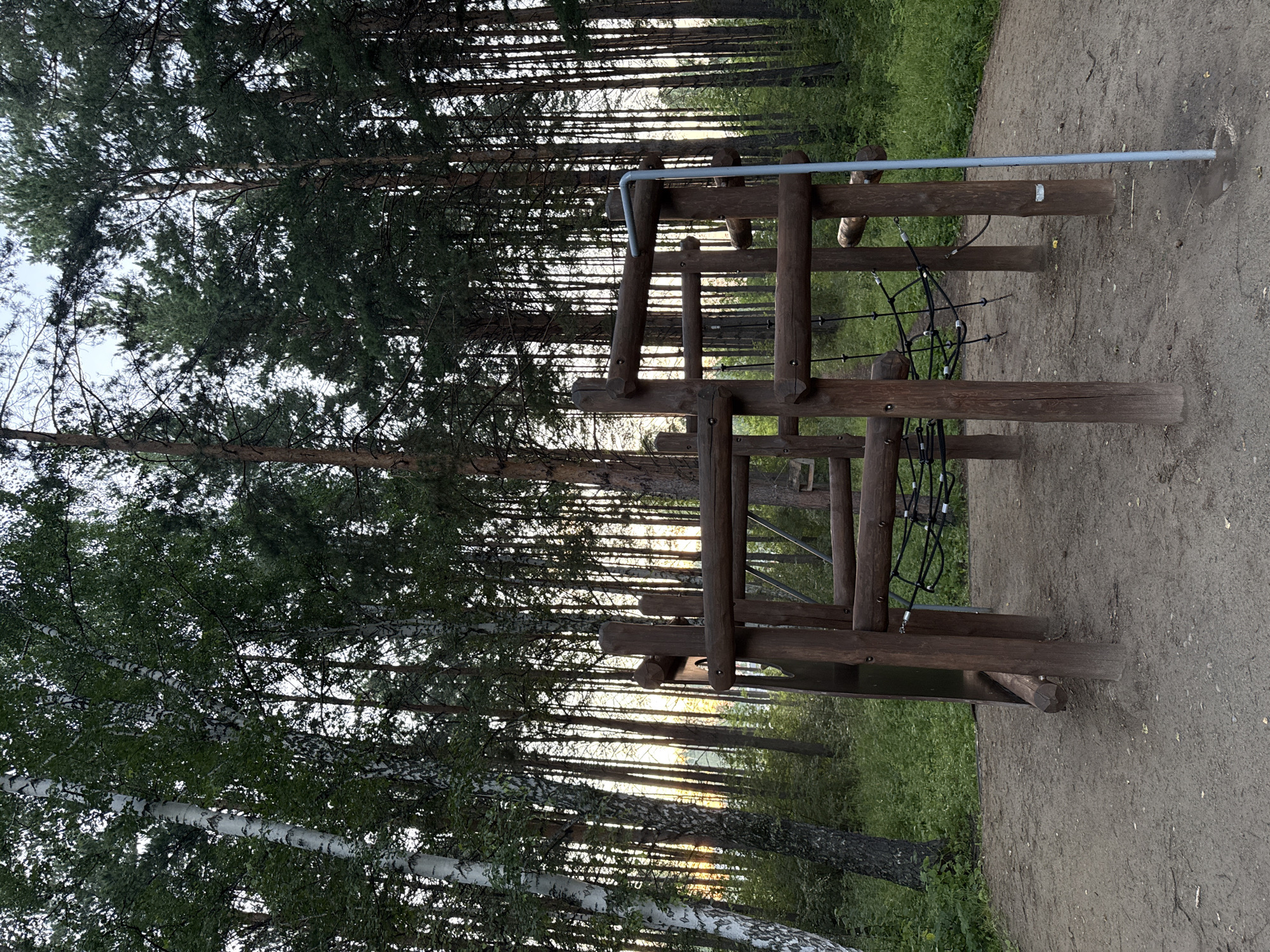 This image shows a wooden playground structure set in a forest clearing. The playground features a series of horizontal wooden beams and vertical posts, with a net suspended between some of the posts. A blue metal pole is visible on the right side of the structure. The ground around the playground is covered in dirt and sand. The background is dominated by tall trees, with a mix of birch and pine trees visible. Sunlight filters through the trees, suggesting it is either early morning or late afternoon. The scene is likely located in a natural park or recreational area within the city of Krasnoyarsk, Russia, given the dense forest environment. There are no people present in the image.
