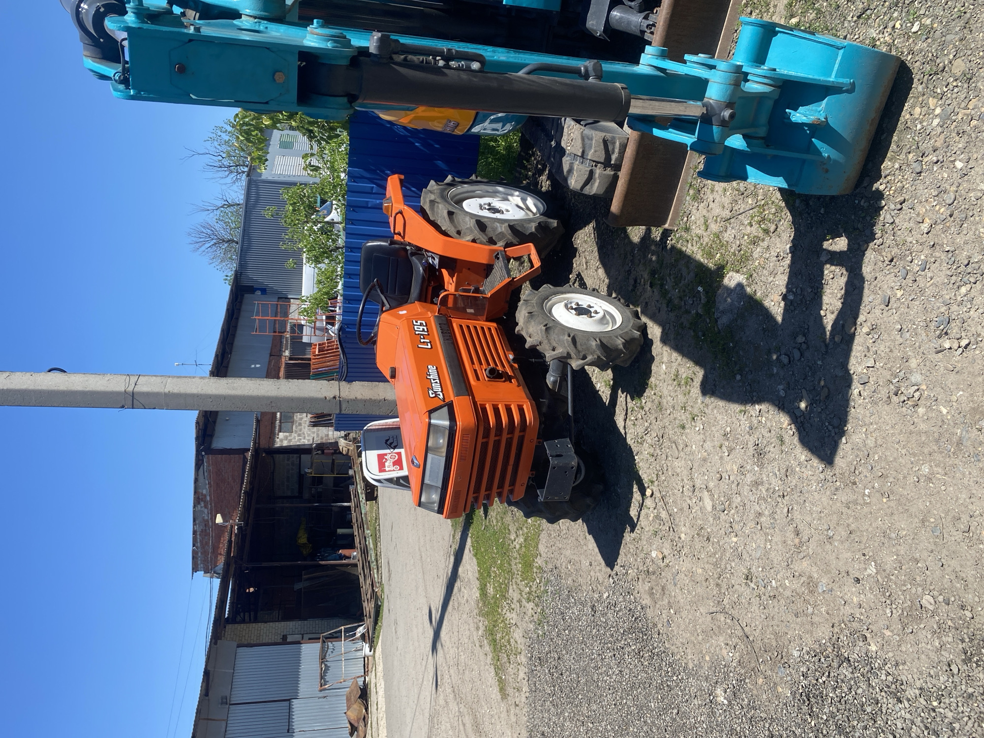 The image depicts a Sunshine LT-195 tractor, colored orange, situated on a gravelly surface. The tractor is partially obscured by a piece of construction equipment, potentially a backhoe, painted in shades of blue. In the background, there is an industrial-looking area with corrugated metal buildings and a concrete utility pole visible against a clear, blue sky. The location is possibly Neftyanik, Russia.