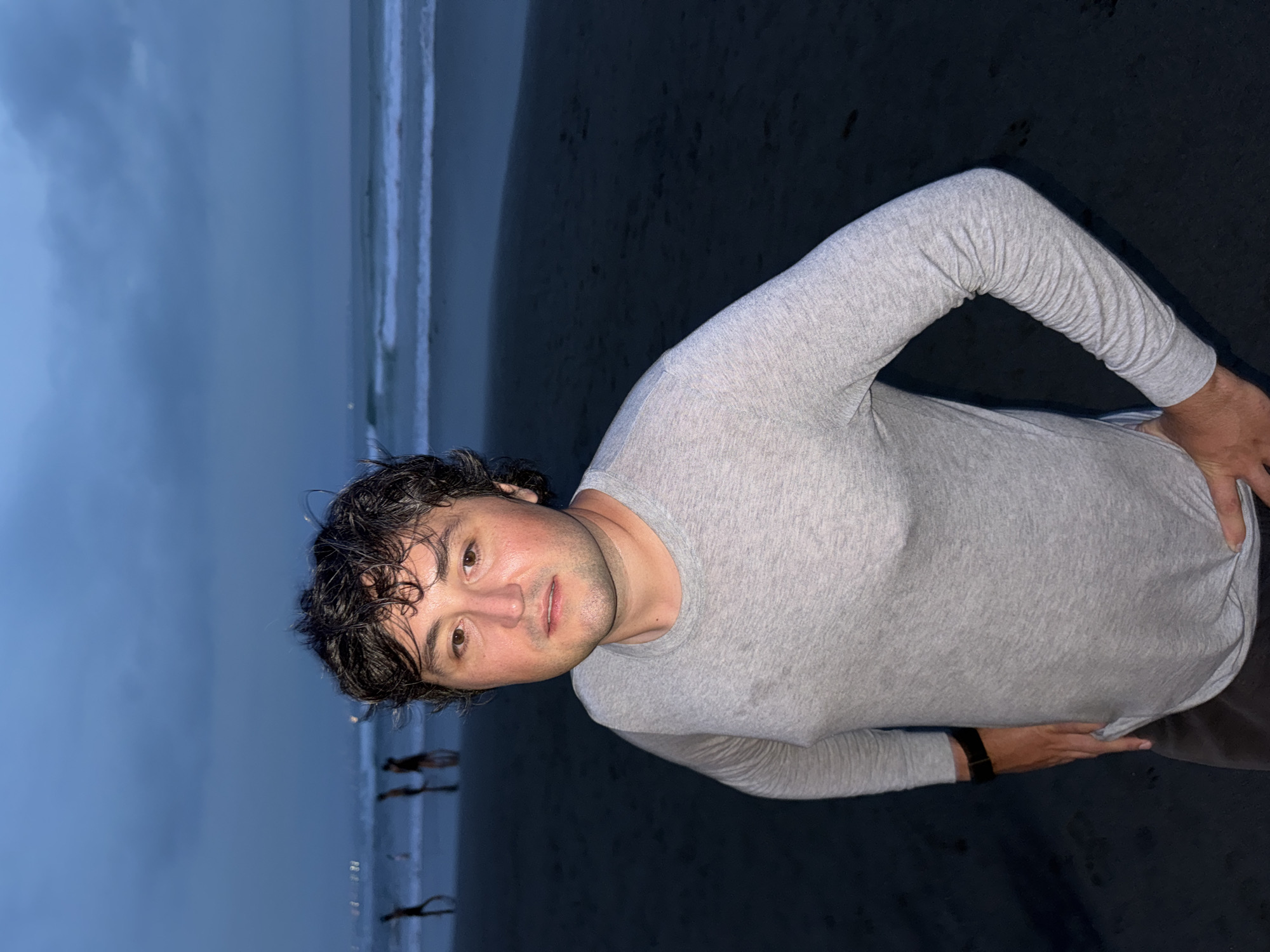 A man stands on a dark sandy beach with ocean waves in the background under an overcast sky, likely at dusk or in the evening. The man is wearing a light gray, long-sleeved shirt and dark pants. He is looking at the camera with a slight smile. His hair is dark and wet, possibly from the sea. Based on the lighting, beach appearance, and vegetation in the background, the location is likely Canggu, Indonesia.