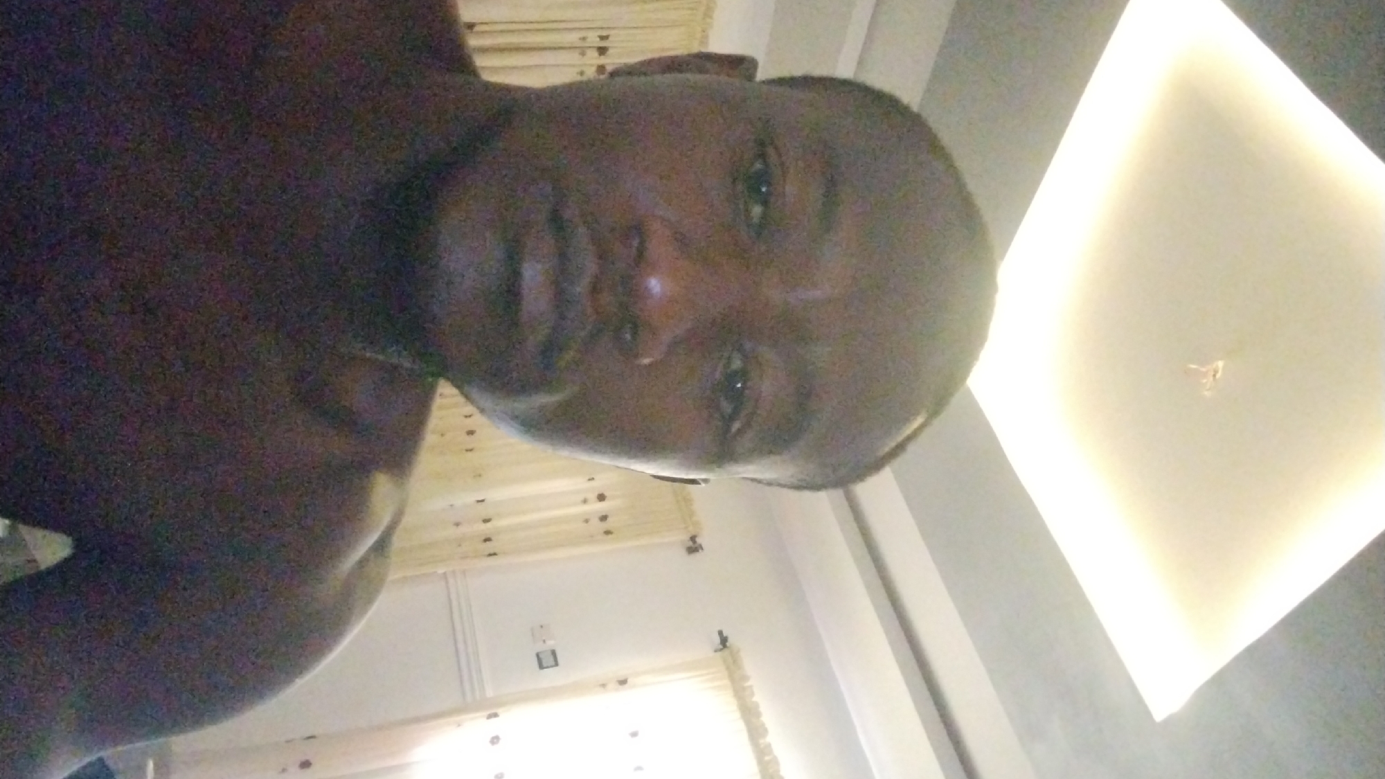 The image shows a dark-skinned, shirtless person's head and upper torso facing the camera. The background includes a window with light-colored curtains, a white wall with a power outlet, and a square recessed light fixture in the ceiling. There is no visible information that specifies the location as Jos, Nigeria.