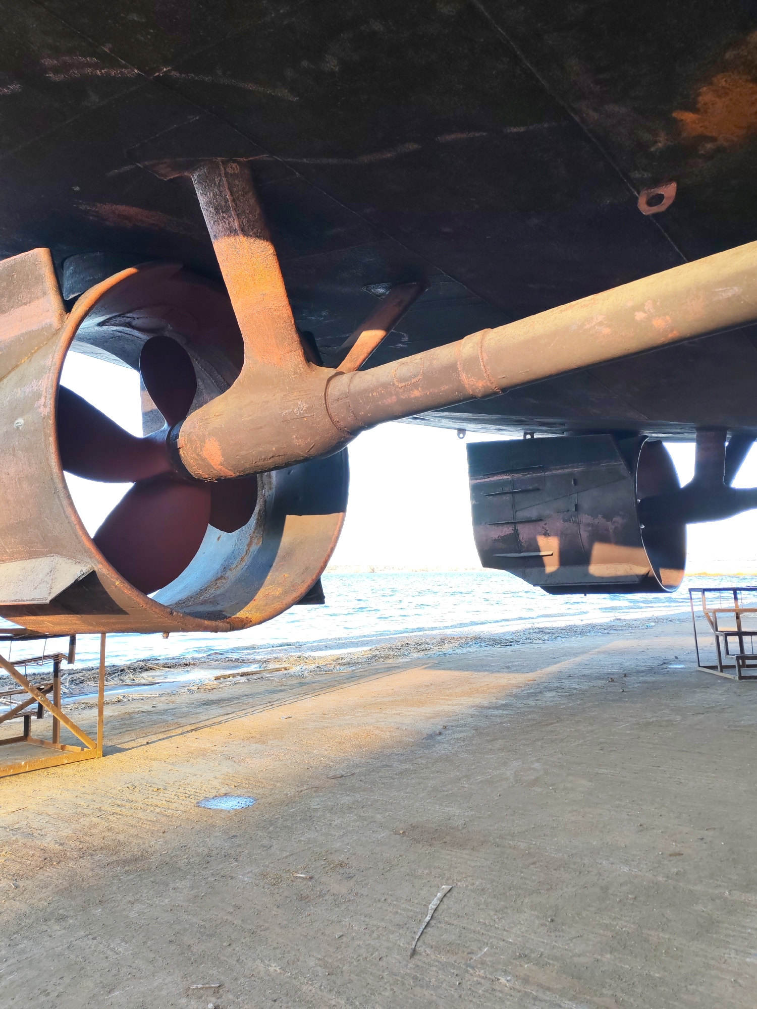 An industrial photograph captures the underside of a large vessel, likely a ship or barge, out of water at a dry dock or slipway in Rybinsk, Russia. The perspective is looking upwards from the ground towards the hull and the water beyond.
The primary subjects are the ship's propulsion system and rudder. On the left, a large, dark metal propeller is visible, featuring three blades and encased within a cylindrical nozzle, commonly known as a Kort nozzle. A robust, rust-colored propeller shaft extends from the hull towards the propeller, supported by a sturdy strut. To the right of the propeller, further along the hull, is a rectangular, dark-colored rudder system. The hull itself appears dark, possibly coated with anti-fouling paint, showing some signs of wear and discolored patches, indicative of time spent in water and exposed to elements. Support structures, made of metal frames, are partially visible beneath the vessel, holding it in place on land.
No people are visible in the image. The scene suggests the vessel is undergoing maintenance, repair, or inspection, given its position out of the water.
The setting includes a flat, greyish-brown ground surface, possibly concrete or compacted earth, which extends into the foreground. A small puddle of water reflects the sky on the ground. In the background, a body of water, likely the Volga River, shimmers under bright sunlight, with a hazy or icy shoreline visible. The time of day appears to be sunny daytime, with strong, directional sunlight casting distinct shadows from the vessel and support structures onto the ground. The overall impression is one of a working shipyard environment. No visible text is present in the image.