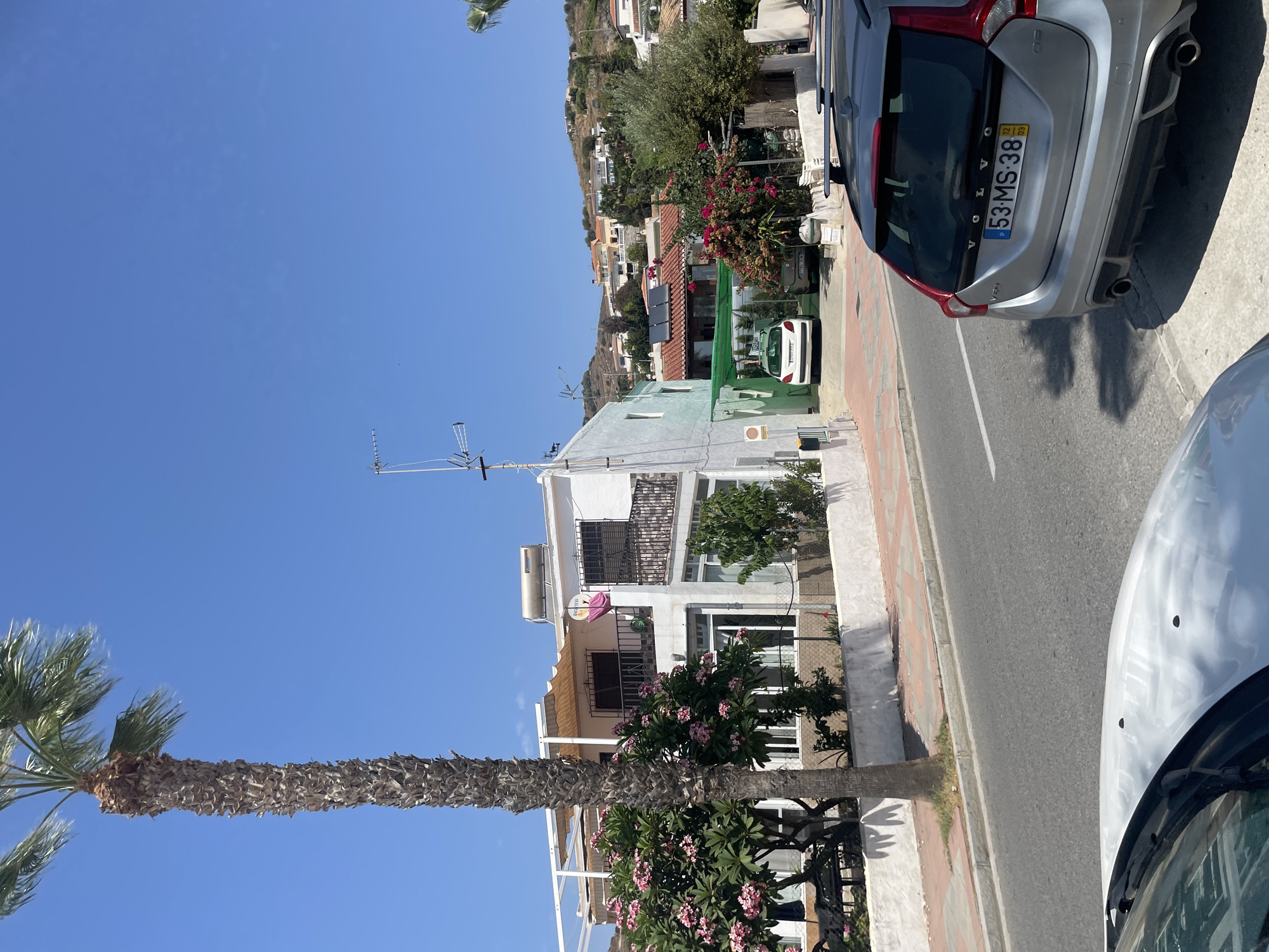 In Almuñécar, Spain, on a sunny day, a silver Volvo V60 is parked on the right side of a two-lane asphalt road. The car's license plate reads "53-MS-38". In the foreground, the hood of a white car is visible. Across the street, a white two-story house with balconies and pink flowering trees in front is situated. Behind the house, a hill covered with residential buildings and vegetation rises. A tall palm tree stands to the left of the house. The sky is clear and blue.