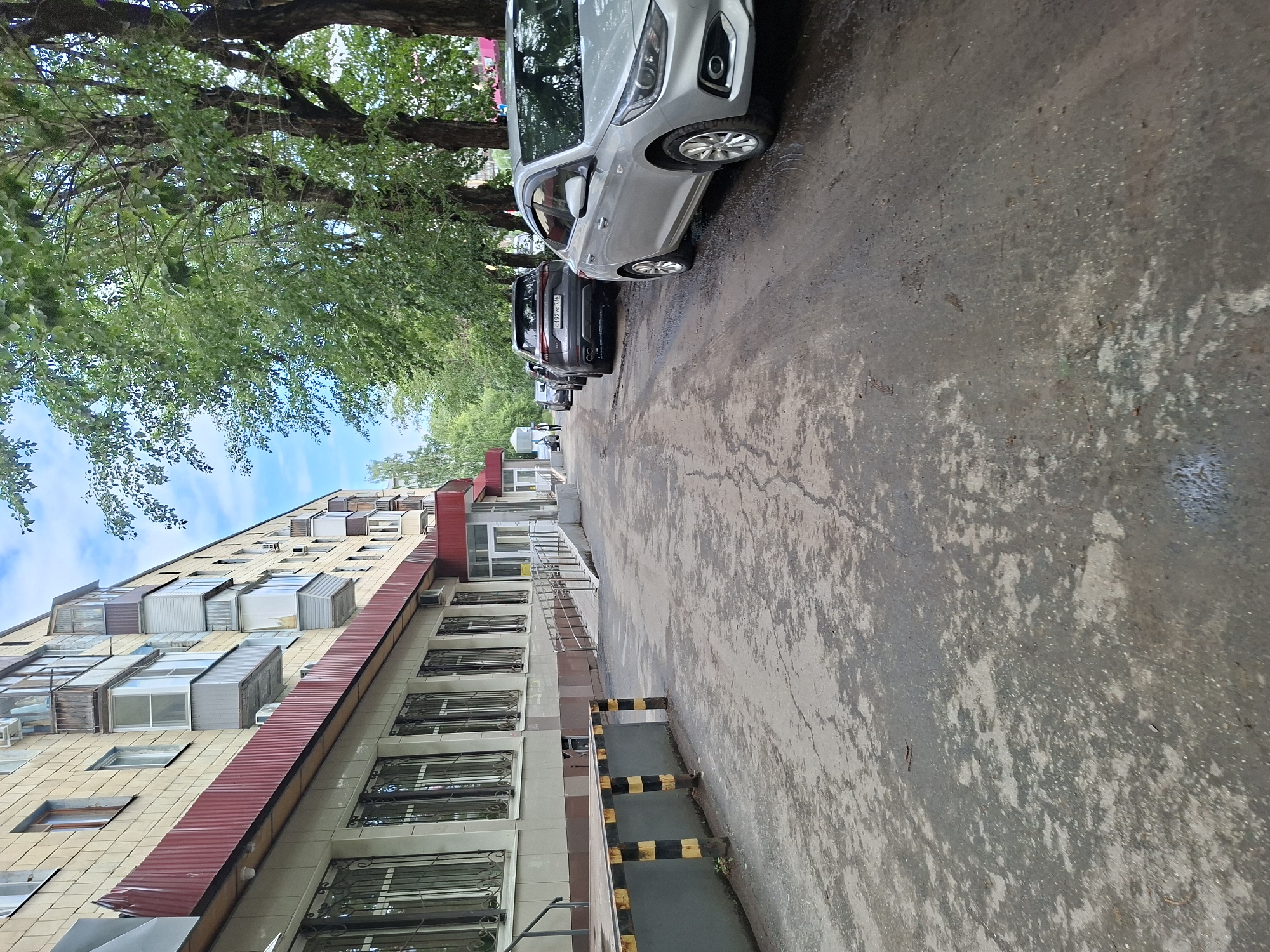 The image depicts an urban street scene in Kazan, Russia, characterized by a multi-story apartment building on the left and parked cars along the right. The apartment building features a beige facade with several balconies and windows. Below the building, there is a red awning. To the right of the building, a ramp leads to an entrance. The street surface appears to be asphalt with some damp patches and debris. Several cars are parked in a row along the right side of the street, including a silver sedan and a dark SUV with visible license plates. Trees with green foliage line the street, casting shadows. The sky is partly cloudy with a bright blue hue. The scene appears to be a typical residential street on a clear day.