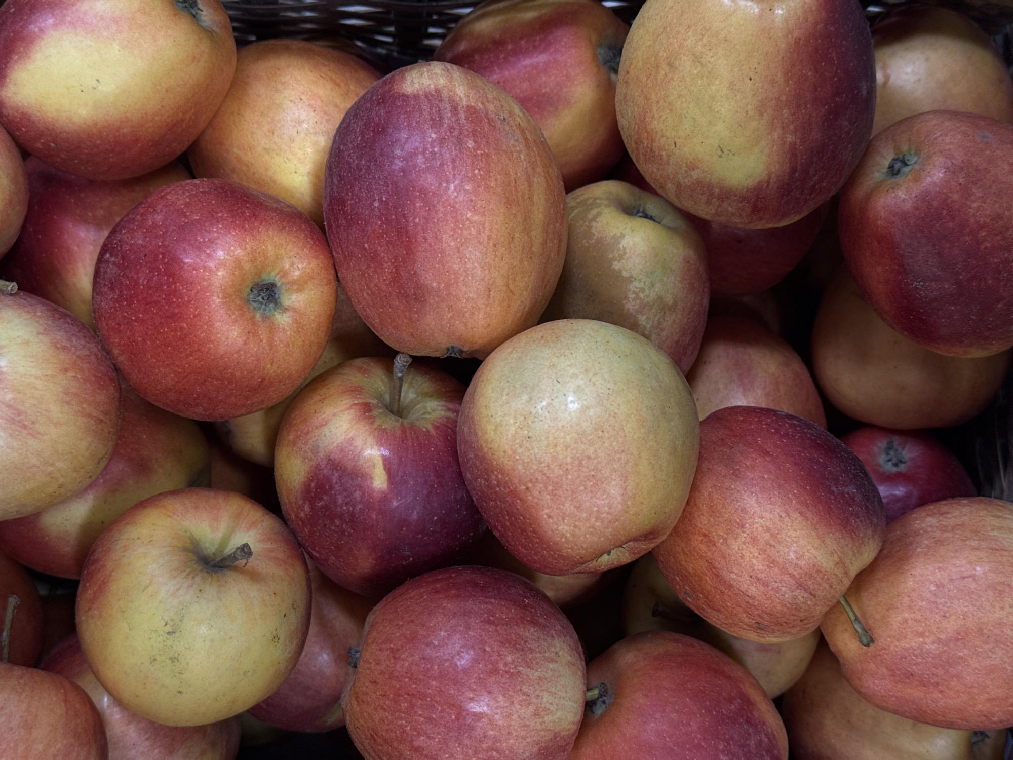 A high-angle, close-up shot shows a basket filled with apples. The apples are mostly red with yellow spots and some are more yellow than red. The apples are tightly packed together. The tops of the basket's wickerwork are visible.