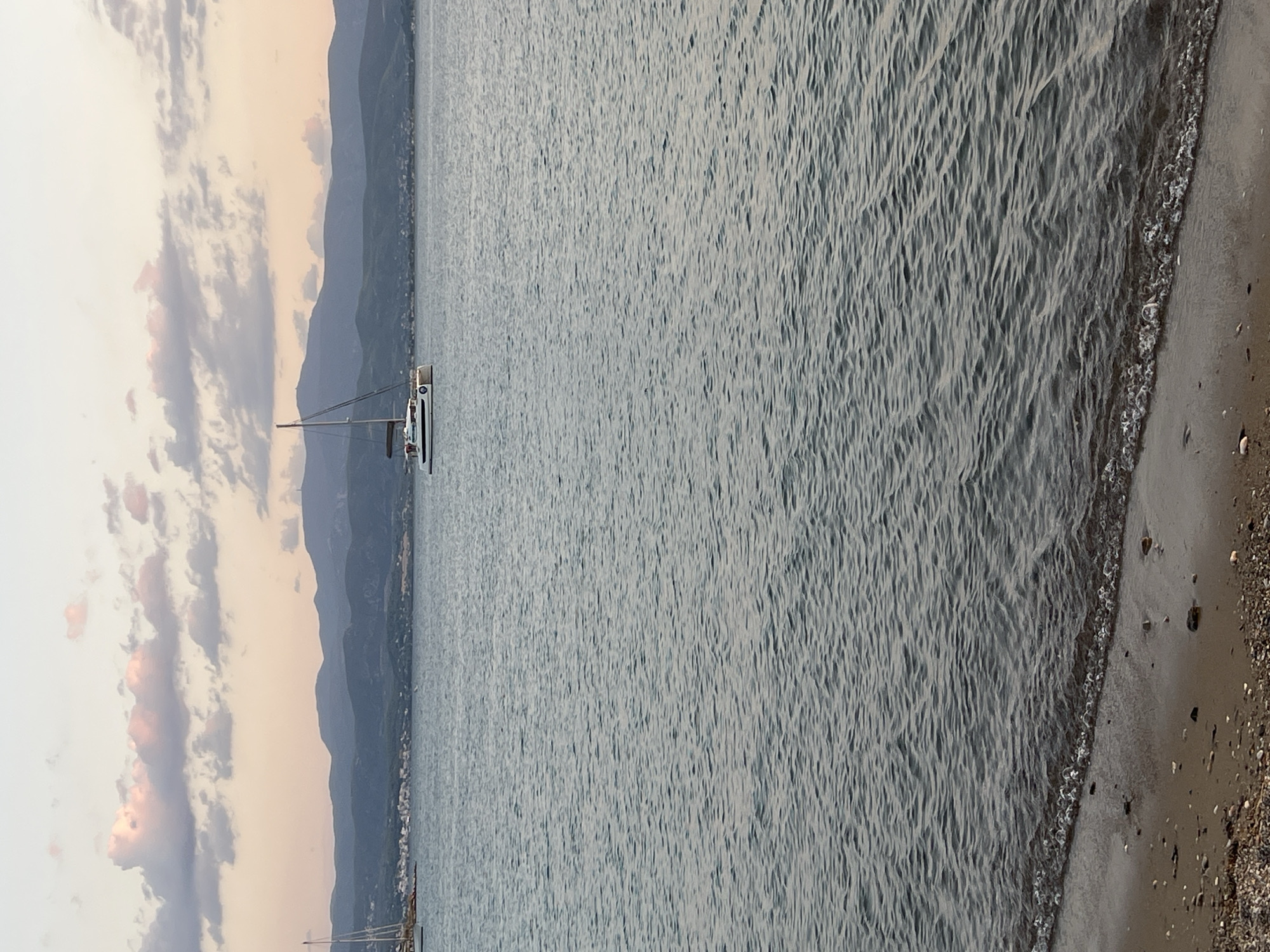 A vertical, eye-level, full shot captures a serene seascape with a sailboat and mountains in the distance. The foreground is a pebble beach with a gentle wave lapping at the shore. The sea is a calm, deep blue, with a slight ripple texture.
In the mid-ground, a white catamaran with a dark sail is anchored, its hull clean and modern. Beyond the sailboat, a town is nestled along the coast, with buildings of varying heights and colors, suggesting a Mediterranean settlement.
The background is dominated by rolling, hazy blue mountains that stretch across the horizon. The sky above is a soft gradient of pastel colors, transitioning from a pale orange near the horizon to a light blue at the top, with scattered, soft pink and gray clouds. The overall atmosphere is tranquil and peaceful, indicative of a late afternoon or early evening.
The scene is likely located in the Eretria Municipal Unit, Greece, given the characteristic landscape of the Aegean Sea with its islands and coastal towns.
