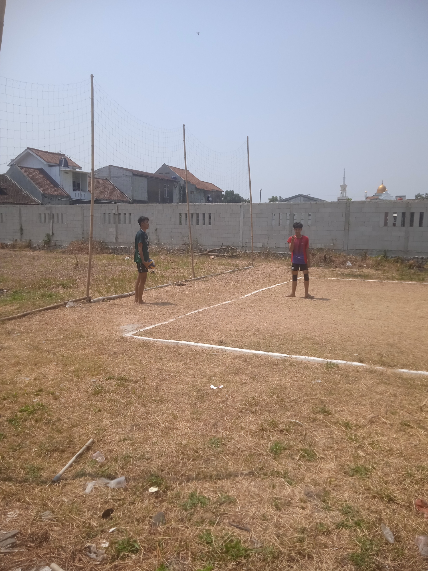 Two young males are depicted on a dry, sandy volleyball court under a clear, bright sky. The court has white lines marking its boundaries. In the background, a chain-link fence stretches across the scene, separating the court from a residential area with houses and a mosque with a golden dome and minaret. The left side of the court has a volleyball net supported by bamboo poles. The male on the left is wearing a dark green t-shirt and dark shorts, holding a volleyball. The male on the right is wearing a red and blue t-shirt and dark shorts, with knee pads, and is in a ready stance. The ground surrounding the court is covered in dry grass and scattered debris, including plastic bags and sticks. The overall impression is an outdoor recreational space in a semi-urban or rural setting in Klodran, Indonesia.