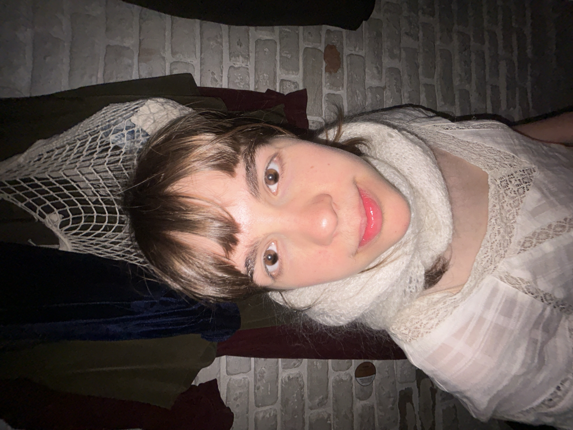 A young individual with brown hair, brown eyes, and fair skin is positioned at an angle in front of a brick wall. They are wearing a white top with lace details and a white scarf. A white net-like object is visible near their head, and clothing items of various colors, including olive green, navy blue, and maroon, are situated beneath them. The location appears to be indoors.