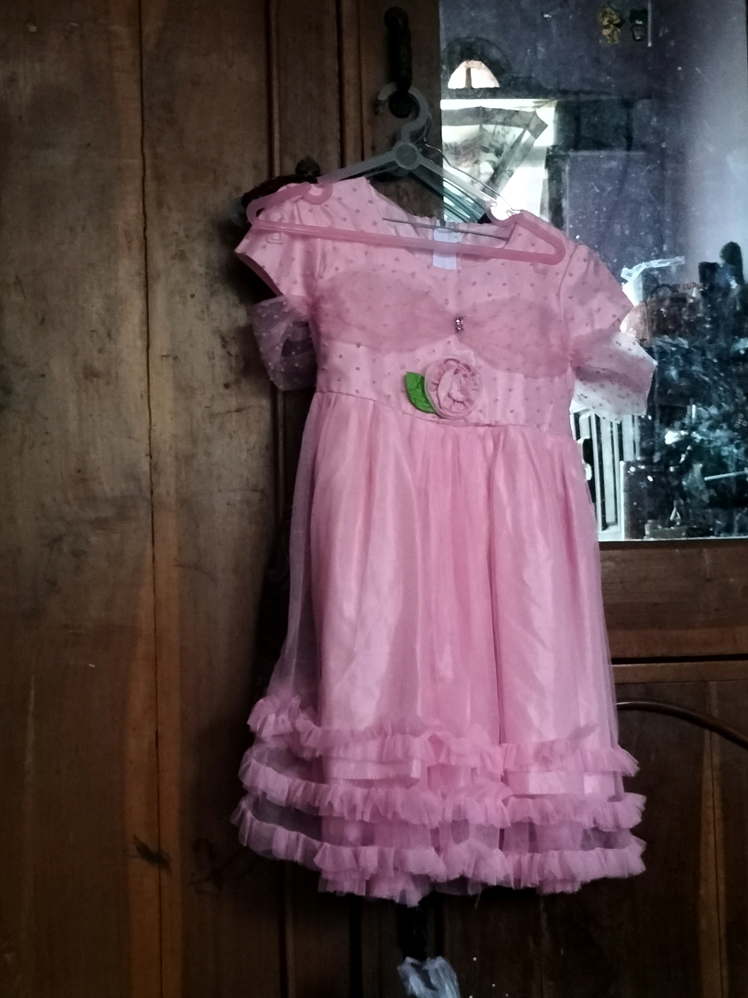 A pink, ruffled, girl's dress hangs on a hanger against a wooden door. The dress is made of sheer fabric with polka dots on the sleeves and bodice, and has a floral applique on the waist. The skirt is layered with ruffles. A mirror is visible in the background, reflecting a window and possibly an outdoor scene. The overall environment appears to be indoors, likely a room in Sambirejo, Indonesia.