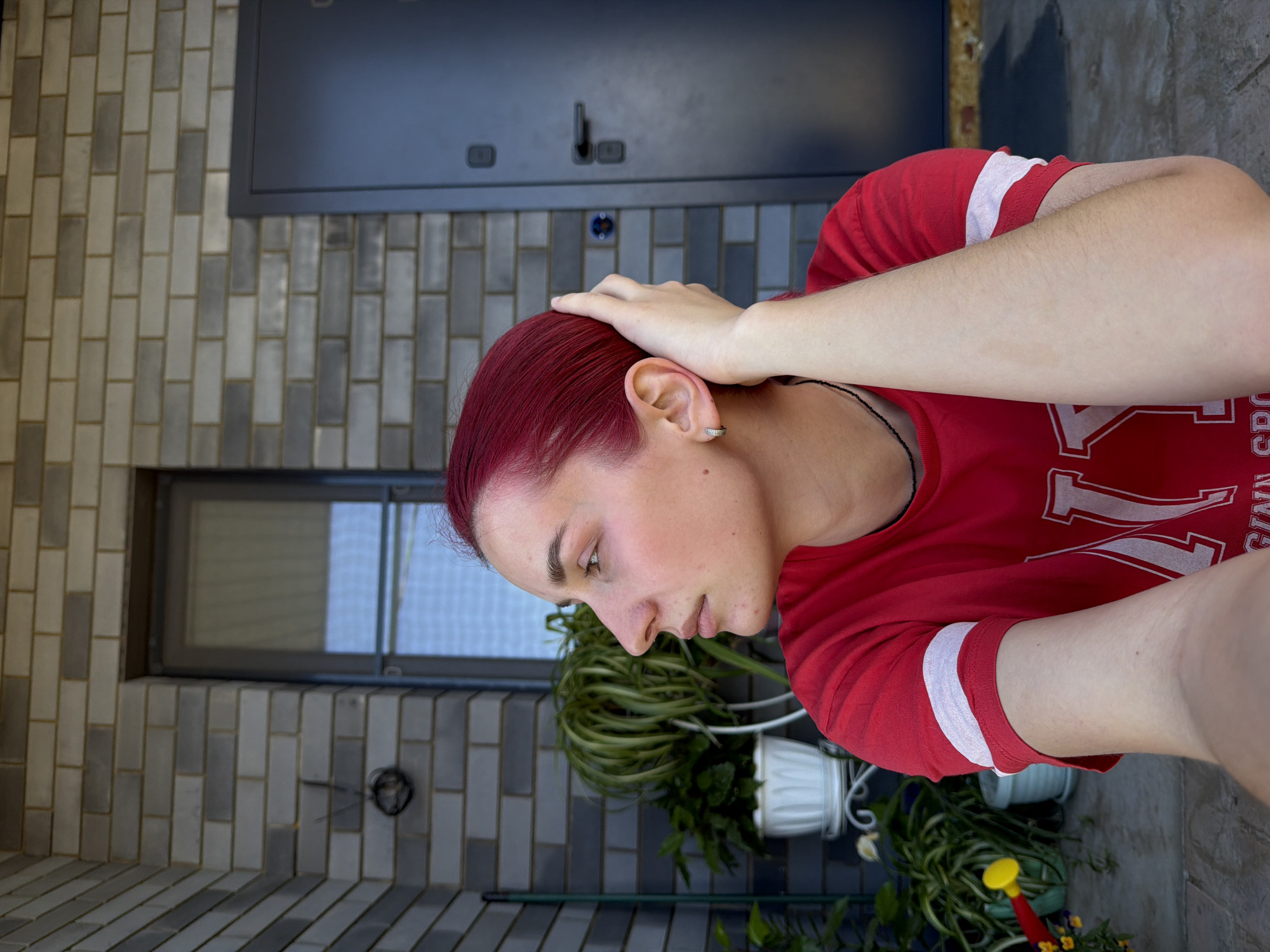 The image depicts a young woman with vibrant red hair sitting outdoors, looking to her left. She is wearing a red t-shirt with white stripes on the sleeves and a white emblem on the chest. Her right hand is raised to her head, touching her hair. In the background, there is a brick wall, a window, a dark-colored door, and several potted plants. The scene appears to be in front of a residential building. The specific location in Chaikovsky, Russia, cannot be determined from the visual information alone.