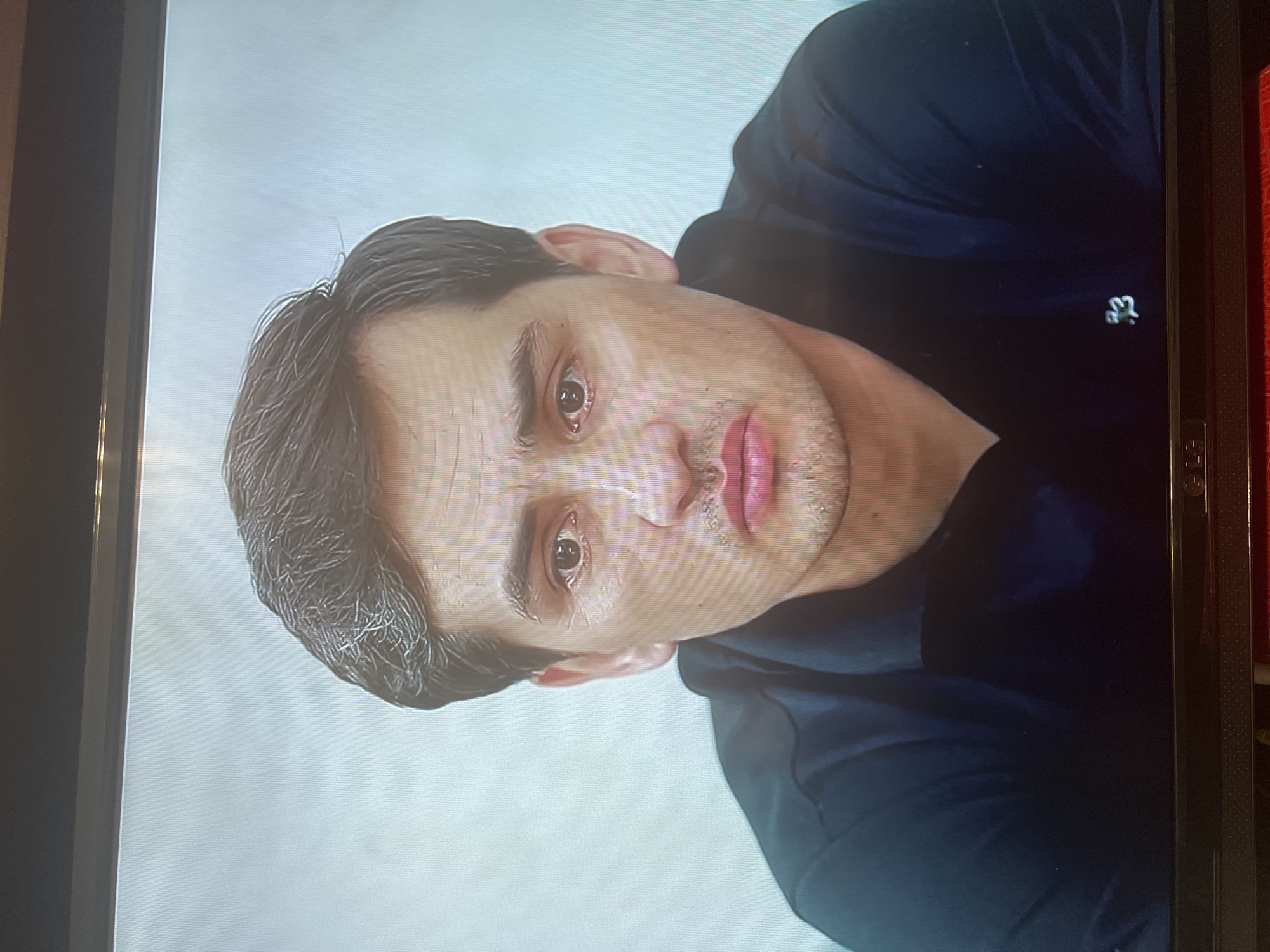 A close-up, head-and-shoulders portrait of a man with dark hair, dark eyes, and light skin. He is wearing a black polo shirt with a small white logo on the left chest. The background is a light, textured color. The man's expression is somber. The image appears to be a still from a video or photograph taken in Osh City, Kyrgyzstan.