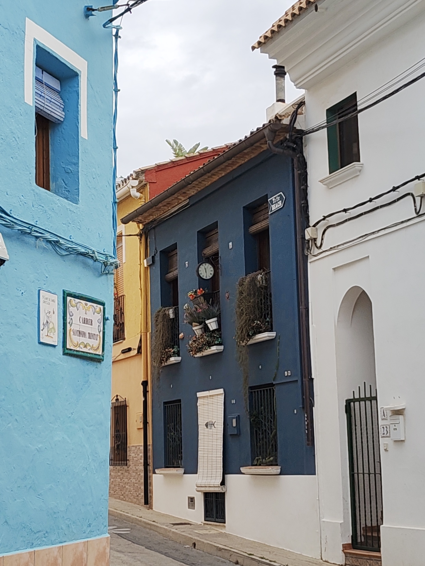 The image captures a narrow street in Dénia, Spain, showcasing colorful buildings tightly packed together. On the left, a blue building features a window with a white frame and a street sign indicating "Carrer Santissima Trinitat." Adjacent to it, a yellow building is partially visible. In the center, a dark blue building displays a clock and flower pots adorning its balconies. A sign pointing "to the Beach" is also visible. On the right, a white building includes a window, an arched doorway, and a gate with the number 23 displayed on a mailbox. Power lines run along the buildings. The street pavement is visible in the foreground, suggesting a quiet residential area.