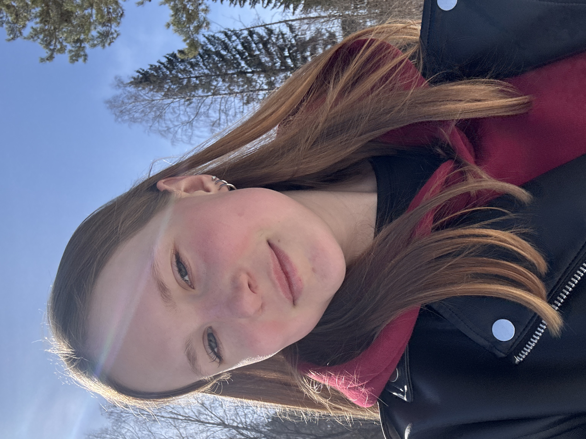 A young woman with light brown hair is pictured in an outdoor setting. Her face is visible and shows rosy cheeks. She is wearing a red hooded sweatshirt under a black leather jacket. The background includes bare trees and a clear blue sky. The location is likely Chaikovsky, Russia, as specified.