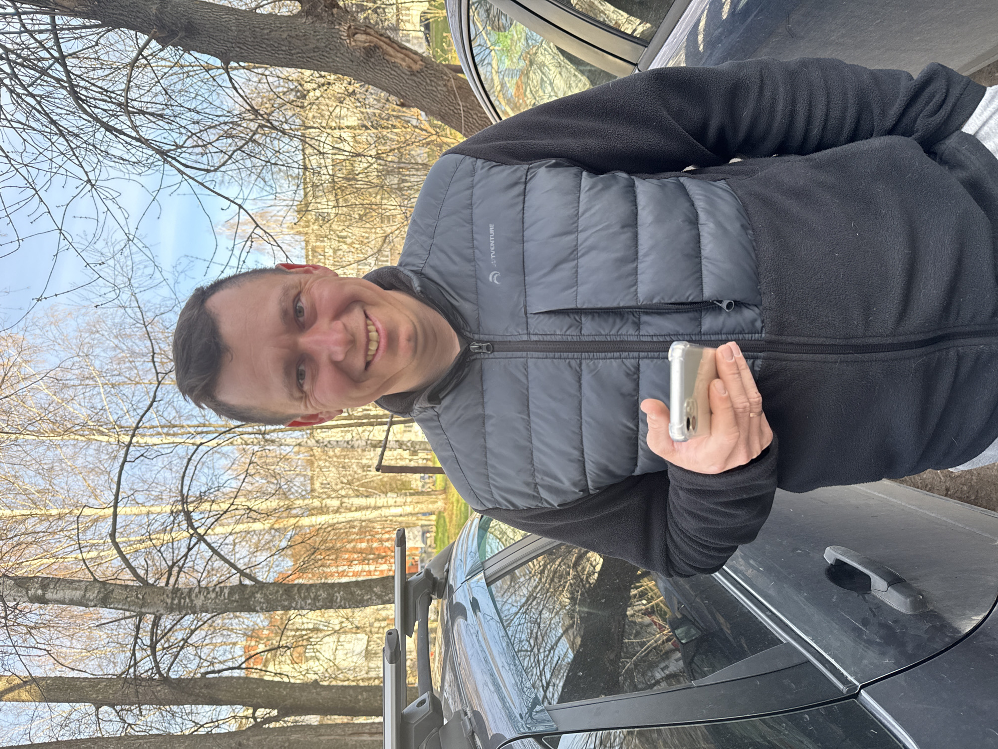 A smiling man is pictured outdoors, standing next to a dark-colored car with a roof rack, against a background of bare deciduous trees and a clear sky. The man, who appears middle-aged with short dark hair, is looking directly at the camera and smiling broadly. He is wearing a black fleece jacket with a gray padded vest over it. The vest has the text "JTT VENTURE" and a crescent moon or C-shaped logo visible on its left chest. He holds a silver-colored smartphone in his right hand.
The bright daylight indicates it is daytime, with clear skies suggesting good weather, though the man's layered clothing implies it might be cool. The bare branches of the trees in the background, along with a faintly visible light-colored building, suggest a season like late autumn, winter, or early spring in Chaikovsky, Russia. The car, partially seen behind the man, reflects the surrounding trees and sky. The presence of a roof rack on the car suggests it might be used for travel or carrying gear.