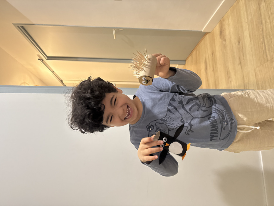 A smiling boy with curly hair holds up two crafts. One is a cardboard tube with toothpicks sticking out, and googly eyes glued on the front. The other is a penguin made from felt. He wears a blue long-sleeved shirt with a dinosaur graphic and tan pants. He stands in a room with white walls and a light-colored wooden floor. A white door is visible in the background.
