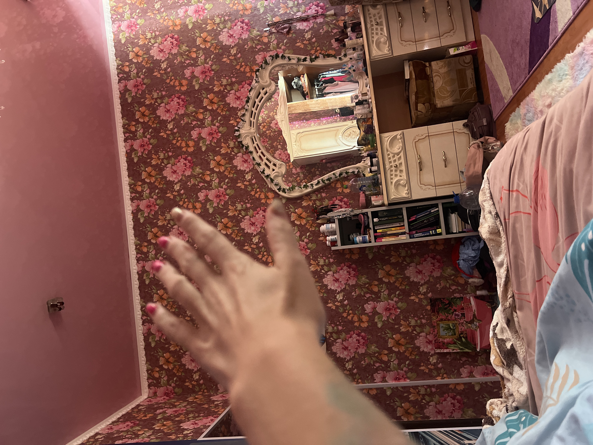 The image depicts a bedroom with floral wallpaper. A hand with pink nail polish is in the foreground, obscuring part of the scene. To the right, a vanity table with a decorative mirror, drawers, and various items on its surface is visible. A bookshelf filled with books and some bottles is located to the left of the vanity. The room also includes a bed with pink and blue bedding. The ceiling is pink. The location is identified as a bedroom in Krasnoe-na-Volge, Russia.
