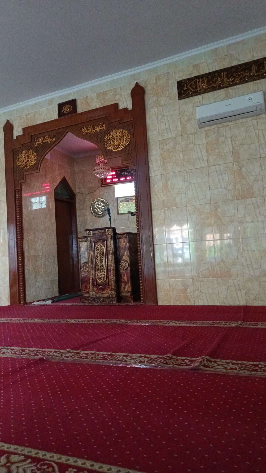 The image depicts the interior of a mosque, likely in Bandung City, Indonesia, featuring a red carpeted floor with decorative golden patterns. In the background, an ornate wooden mihrab (prayer niche) is visible, adorned with Arabic calligraphy. Inside the mihrab, a wooden minbar (pulpit) stands, also decorated with intricate designs. A microphone is positioned on the minbar. Above the minbar, a digital display shows time and text, with the word "NURUL JA" prominently displayed, potentially indicating the mosque's name or an event. A chandelier hangs from the ceiling, casting a pinkish light. The walls are tiled with a light-colored marble pattern. An air conditioning unit is mounted on the wall to the right of the mihrab. The overall ambiance suggests a place of worship, with an emphasis on Islamic art and architecture.