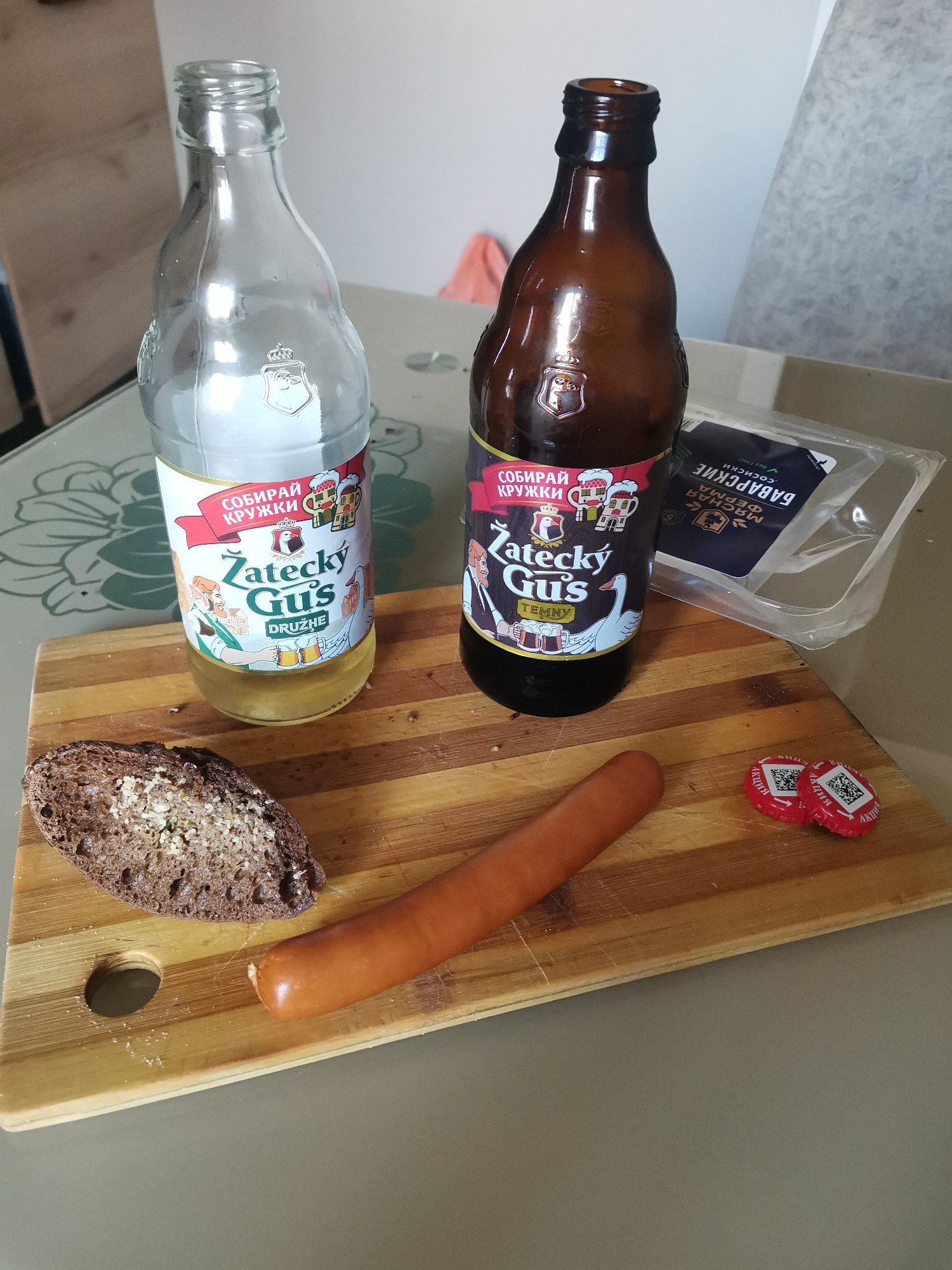 A still life scene on a table, featuring two bottles of "Zatecky Gus" beer, one clear glass and one brown glass, a piece of dark bread with seeds and seasoning, a single frankfurter, two red bottle caps with QR codes, and a plastic package of sausages in the background. The bottles are labeled with colorful graphics depicting characters and beer mugs. The bread and frankfurter are placed on a wooden cutting board. The background shows a blurred interior setting with furniture. The image does not provide any specific details indicating the location in the city of Chaikovsky, Russia.