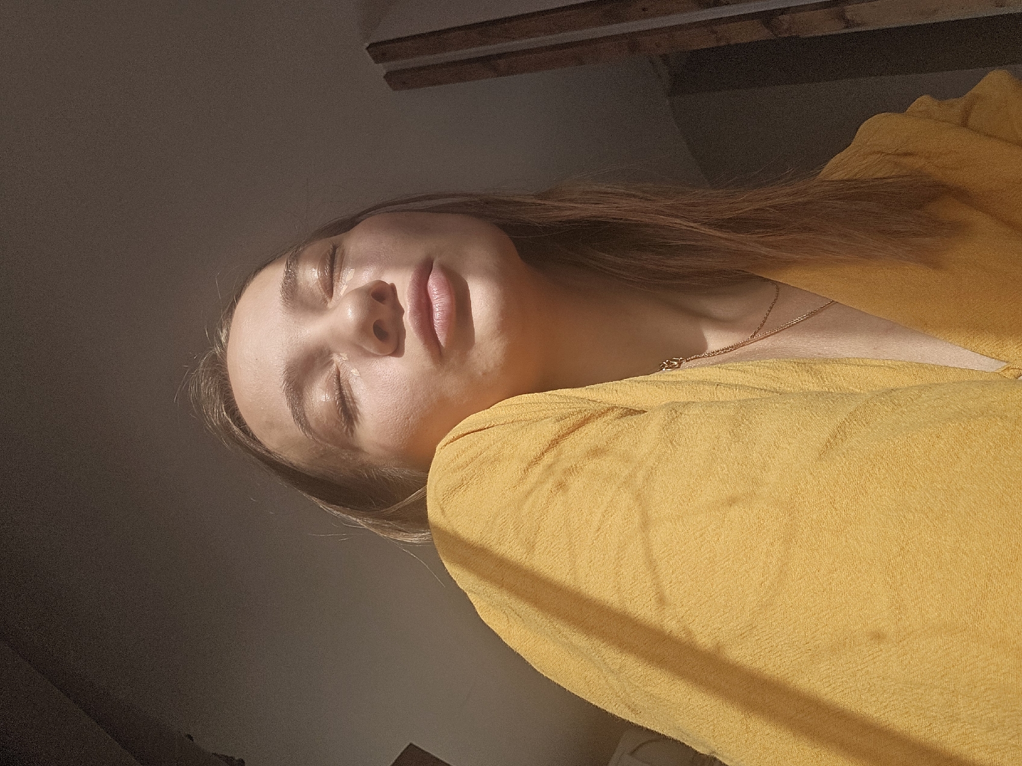A portrait of a young woman with her eyes closed, her head tilted back and to the side. She is wearing a yellow top and a delicate gold necklace. Sunlight illuminates her face, casting a warm glow on her skin and highlighting the natural curve of her lips. Her hair, a light brown, falls around her shoulders and onto the yellow fabric of her clothing. The background is dimly lit, with a hint of wooden structure visible at the top of the frame. The lighting suggests it is either early morning or late afternoon, with the sun casting long shadows. The woman's expression appears peaceful and serene, as if she is enjoying the warmth of the sun. No text is visible in the image.
