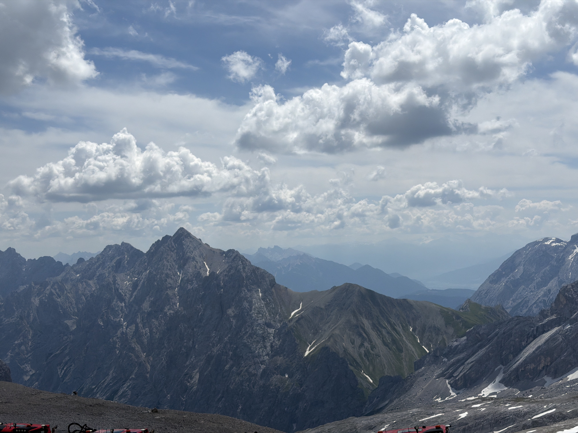 The image captures a panoramic view of a mountainous landscape, likely from a high vantage point in or near Garmisch-Partenkirchen, Germany. The foreground features a rocky, barren terrain, with remnants of what appear to be red recreational vehicles or equipment. Beyond this, a series of imposing, jagged mountain ranges dominate the scene, their rugged surfaces displaying variations in rock texture and color. Patches of snow are visible on the higher slopes, suggesting an alpine environment. In the distance, successive layers of mountains fade into a hazy blue, creating a sense of depth and vastness. The sky above is a mixture of blue and white, with large, fluffy cumulus clouds casting shadows on the mountains below. The overall impression is one of dramatic natural beauty and rugged, untamed terrain.