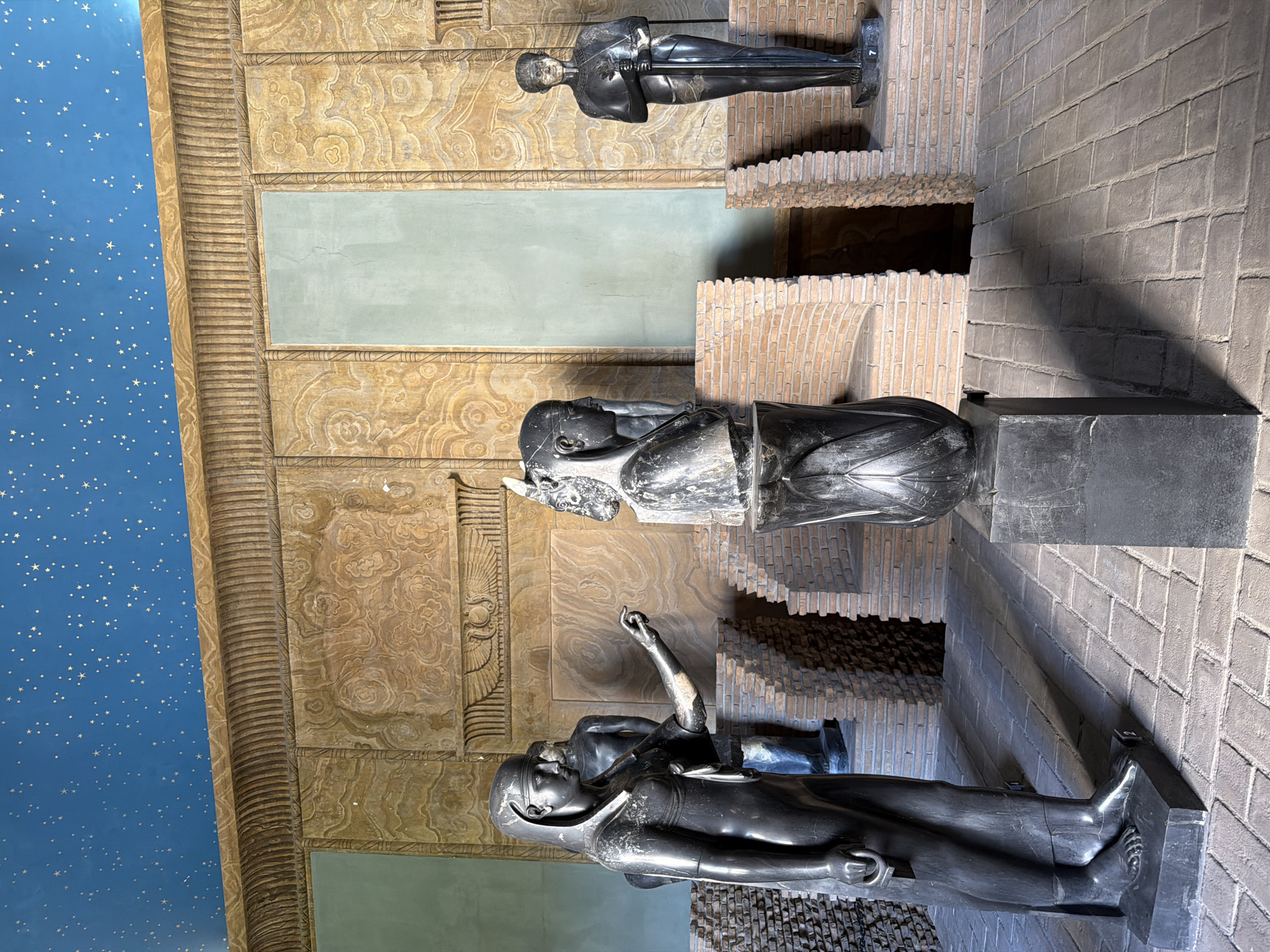 The image shows an interior exhibition space with several dark stone statues. In the foreground, a female statue with a headdress and a long dress stands on a pedestal, pointing her right hand towards a bust of a woman with a similar headdress. Behind them, another male statue stands holding a sword. The walls are decorated with textured panels and features two rectangular green panels. The ceiling is painted blue with scattered gold stars. The floor is paved with rectangular bricks. In the background, there are curved brick walls forming alcoves, and another male statue holding a sword. The overall setting appears to be a museum or exhibition hall, likely an ancient Egyptian-themed display. The presence of "Vatican City, Vatican City" in the prompt suggests this location is within the Vatican Museums.