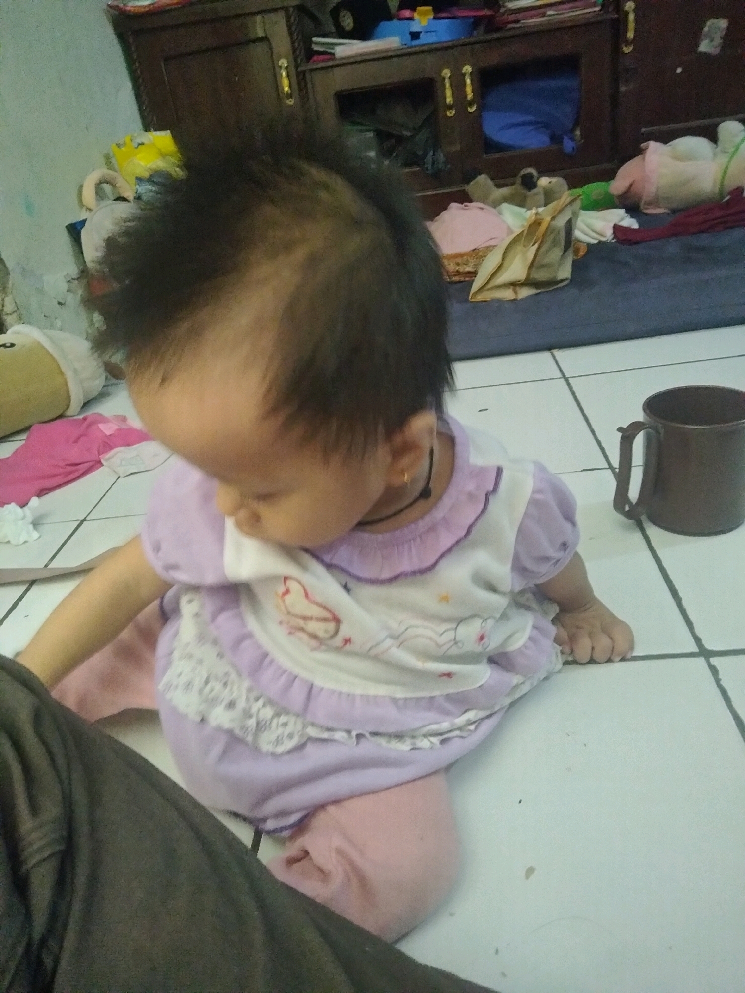 In a room in Suka Sari, Indonesia, a baby with dark hair is sitting on a tiled floor. The baby is wearing a purple and white outfit with a ruffled collar and lace trim. A dark brown mug is on the floor to the right of the baby. In the background, a wooden cabinet is visible, along with various stuffed animals and blankets scattered on the floor. The lighting suggests it may be daytime, and the overall scene appears to be a residential indoor space.