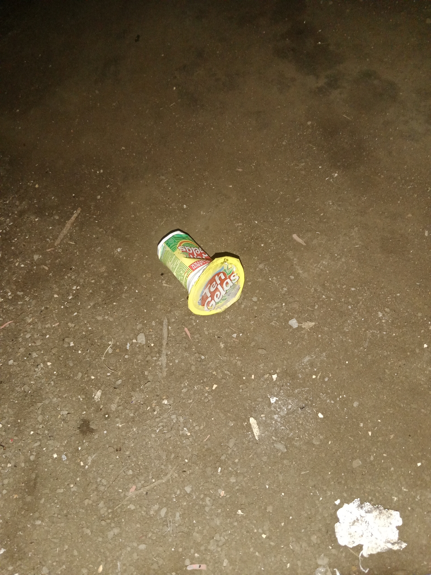 A single, discarded plastic cup of "Teh Gelas" brand iced tea lies on a granular, dirt-like surface. The cup is yellow with green and white branding. It is lying on its side, with the lid partially peeled back, revealing the empty interior. The surface it rests on appears to be unpaved ground, possibly a roadside or undeveloped area, with small stones and debris scattered about. The lighting suggests it is either nighttime or a dimly lit indoor space. The environment appears to be the exterior of a building, or an outdoor area within the village of Ringinrejo, Indonesia, characterized by its rough, unpaved terrain and sparse debris. No individuals are visible in the image, nor are there any signs of recent activity or interaction. The scene is static and suggests neglect or casual littering.