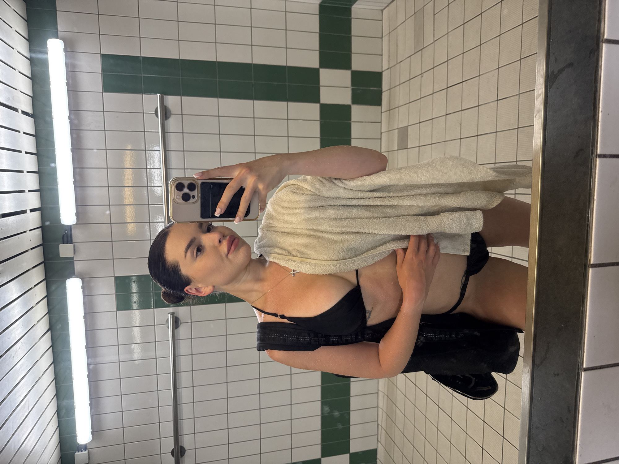 A young woman, with her hair in a bun, takes a selfie in a tiled locker room. She is wearing a black bikini and holding a cream-colored towel. She has a black bag slung over her shoulder. The locker room features white tiled walls with green accents, fluorescent lighting, and a metal rack. The floor is also tiled with a grid pattern. A large mirror reflects the scene. The image is a medium shot, focusing on the woman and her reflection. The location is not specified in the image, but the request states it is the city of Prague, Czechia.