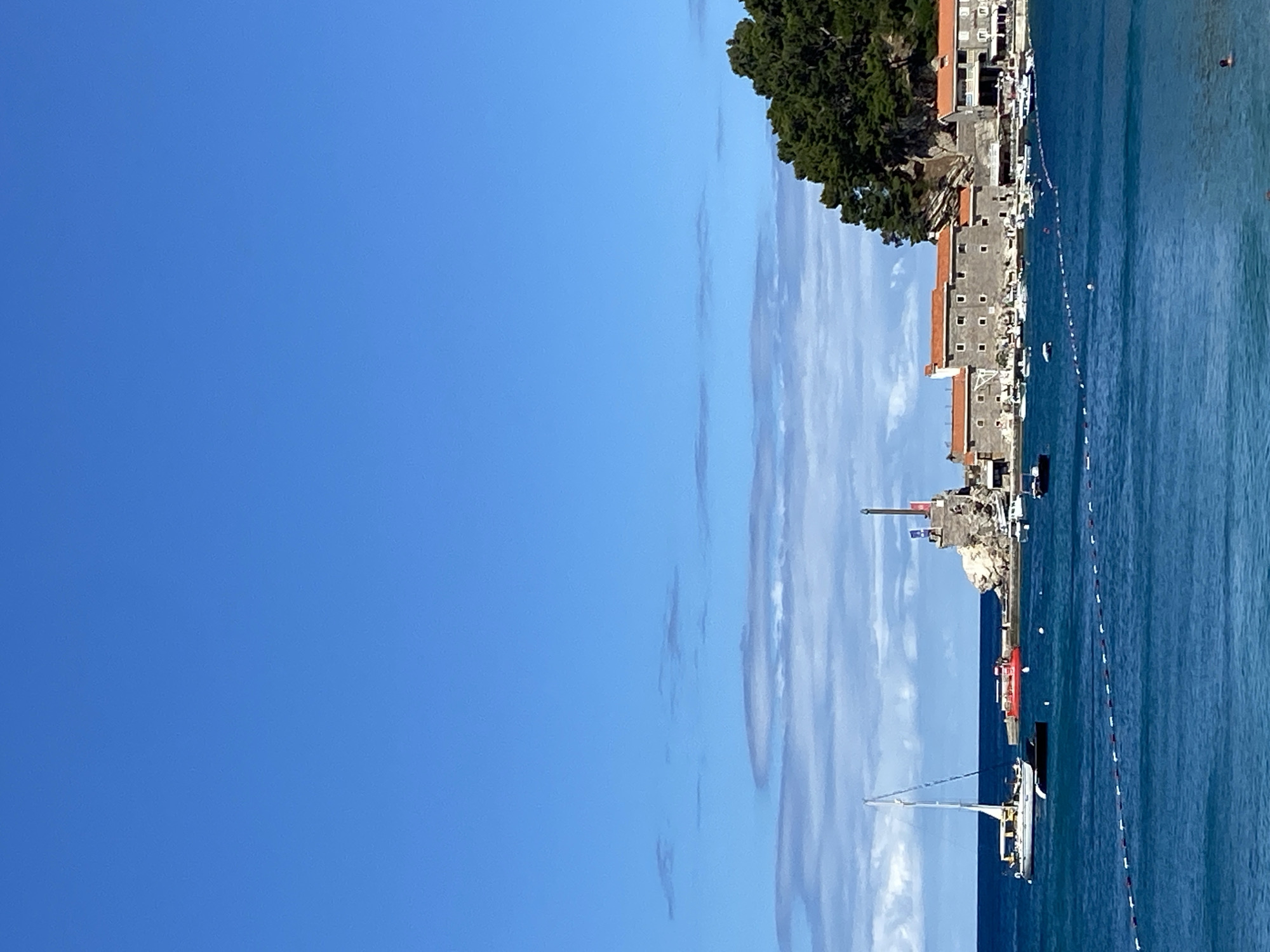 The image shows a view of Petrovac centar, Montenegro, on a clear day. The sky is a vibrant blue with a few wispy clouds. In the foreground, the deep blue sea stretches out, with a white and red buoy line marking a section of the water. A few boats are visible on the water, including a white sailboat with its sail up on the left side of the image.
In the middle ground, the coastline features a cluster of stone buildings with red-tiled roofs. A small rocky island juts out from the shore, topped with a stone structure that appears to be a lighthouse or tower, adorned with flags. Several smaller boats are docked or anchored near the buildings.
To the right of the buildings, a steep, tree-covered hill slopes down towards the sea. The overall scene depicts a picturesque coastal town with a tranquil atmosphere.