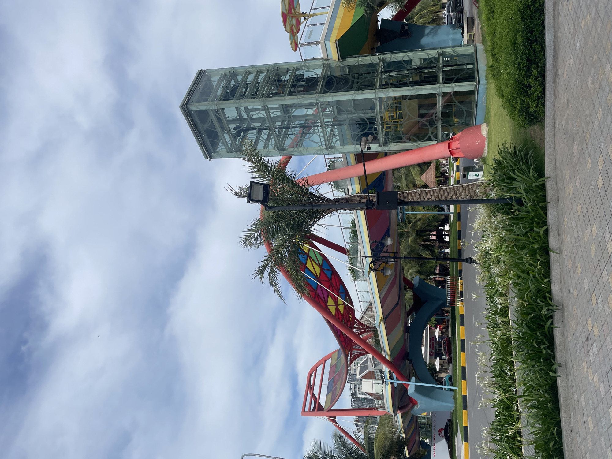 This image depicts a vibrant pedestrian bridge with a distinctive red, curved structure, adorned with colorful panels in shades of yellow, green, and blue. Adjacent to the bridge is a modern glass elevator tower, offering views of the surrounding structures and foliage. In the foreground, a neatly manicured lawn is planted with lush green bushes and delicate white flowers, leading to a paved walkway. A street with a yellow and black striped median separates the walkway from the bridge area. Palm trees are scattered throughout the scene, adding to the tropical ambiance. The sky above is overcast with scattered clouds. The scene appears to be set in a public space, likely a resort or entertainment area, given the whimsical design of the bridge and the presence of a resort-like structure in the background. While no individuals are clearly visible or identifiable, the environment suggests a public gathering space or a tourist destination.