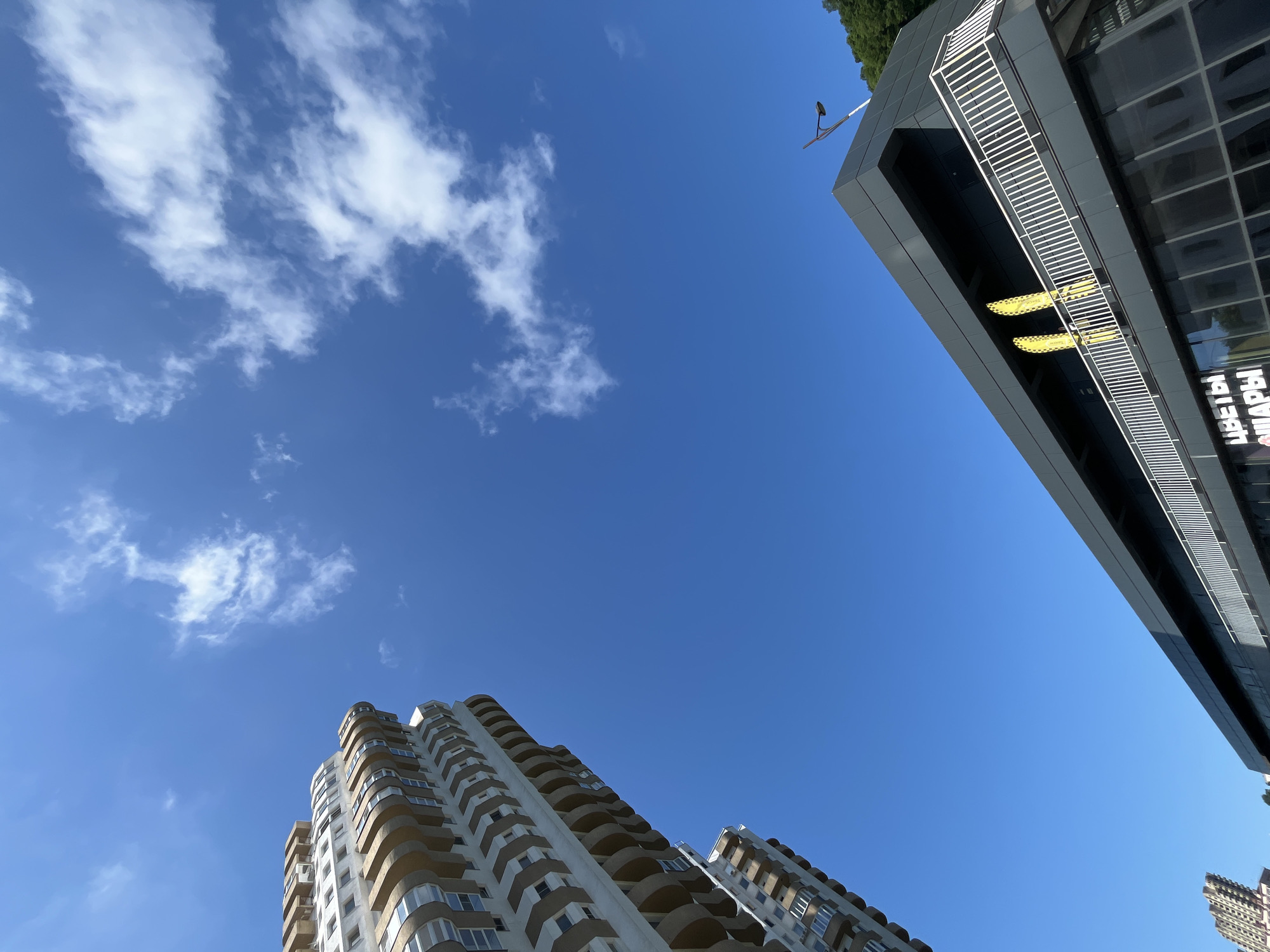 This is a low-angle, wide shot of a clear blue sky with scattered white clouds, taken from a city environment in Sochi, Russia. Two modern buildings are visible. On the right, a dark, multi-story building with large glass windows and white horizontal accents along a balcony area dominates the upper right quadrant. Two yellow signs with black text are visible on this building. The text is partially obscured and appears to be in Cyrillic, with one sign reading "ТАЙМ" and the other "ТАЙМ.". On the left and bottom of the frame, a light-colored, multi-story residential building with curved balconies and numerous windows is visible. The building has a distinctive architectural style with repeated wave-like patterns along its facade. A street lamp is visible on top of the dark building, and a small section of green foliage is visible at the very top right of the frame. The bright sky and clear visibility suggest it is daytime, likely mid-morning or afternoon, with pleasant weather. The overall mood is serene and upward-looking, focusing on the contrast between the man-made structures and the expansive sky.