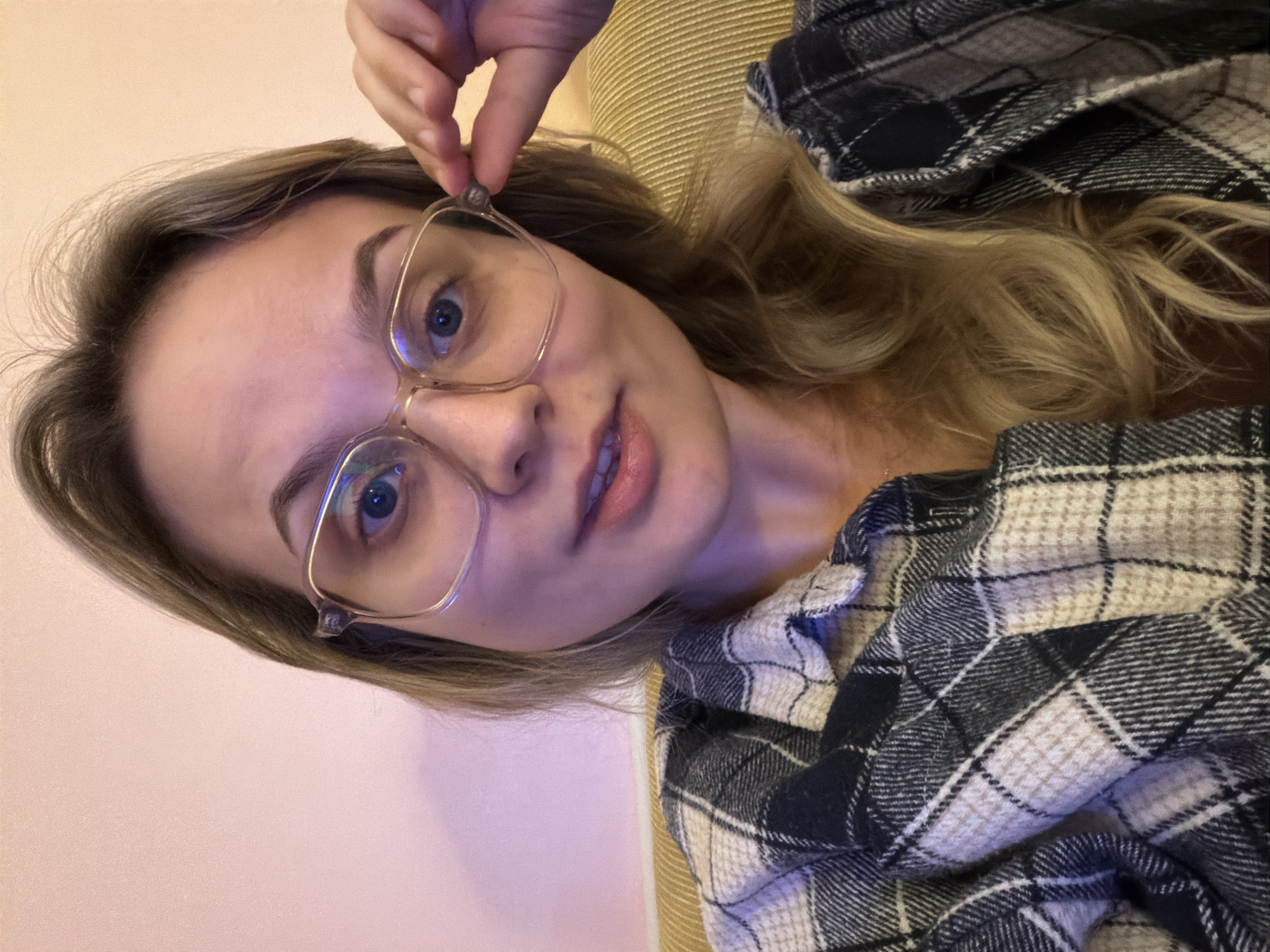 A front-facing portrait depicts a female individual with light brown hair, blue eyes, and light skin, wearing large, clear-framed eyeglasses. Her lips are slightly parted. She is dressed in a black, white, and tan plaid shirt. Her left hand is raised, gripping the right temple of her eyeglasses. The background features a plain, light pink wall. A portion of a yellowish-gold horizontally striped piece of furniture is visible to her right, suggesting she is seated or leaning. The scene is indoors. The specific city location of Tel-Aviv, Israel, cannot be determined from the visual elements within the image.