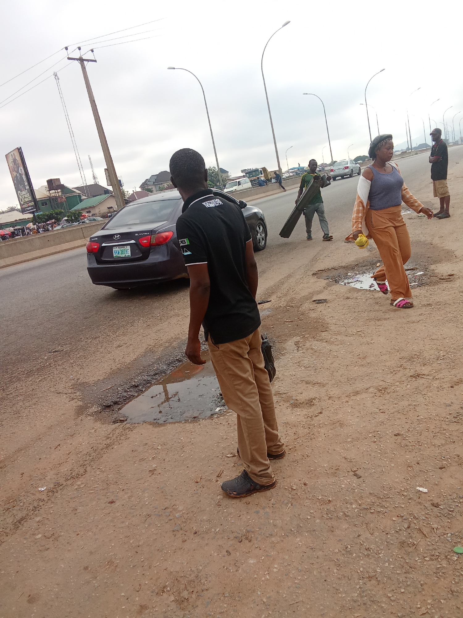 A medium shot shows a group of people on a dusty, unpaved road next to a multi-lane highway. A dark gray Hyundai Elantra is parked on the highway in the background. A man wearing a black t-shirt and brown pants stands in the foreground, facing away from the camera. To his right, a woman in tan pants and a blue top walks with a yellow object in her hand. To her left, a man in a green shirt carries a long, flat object. In the distance, more people and vehicles are visible. The sky is overcast. The scene appears to be in Kagini, Nigeria, indicated by street signs in the background and the overall environment.