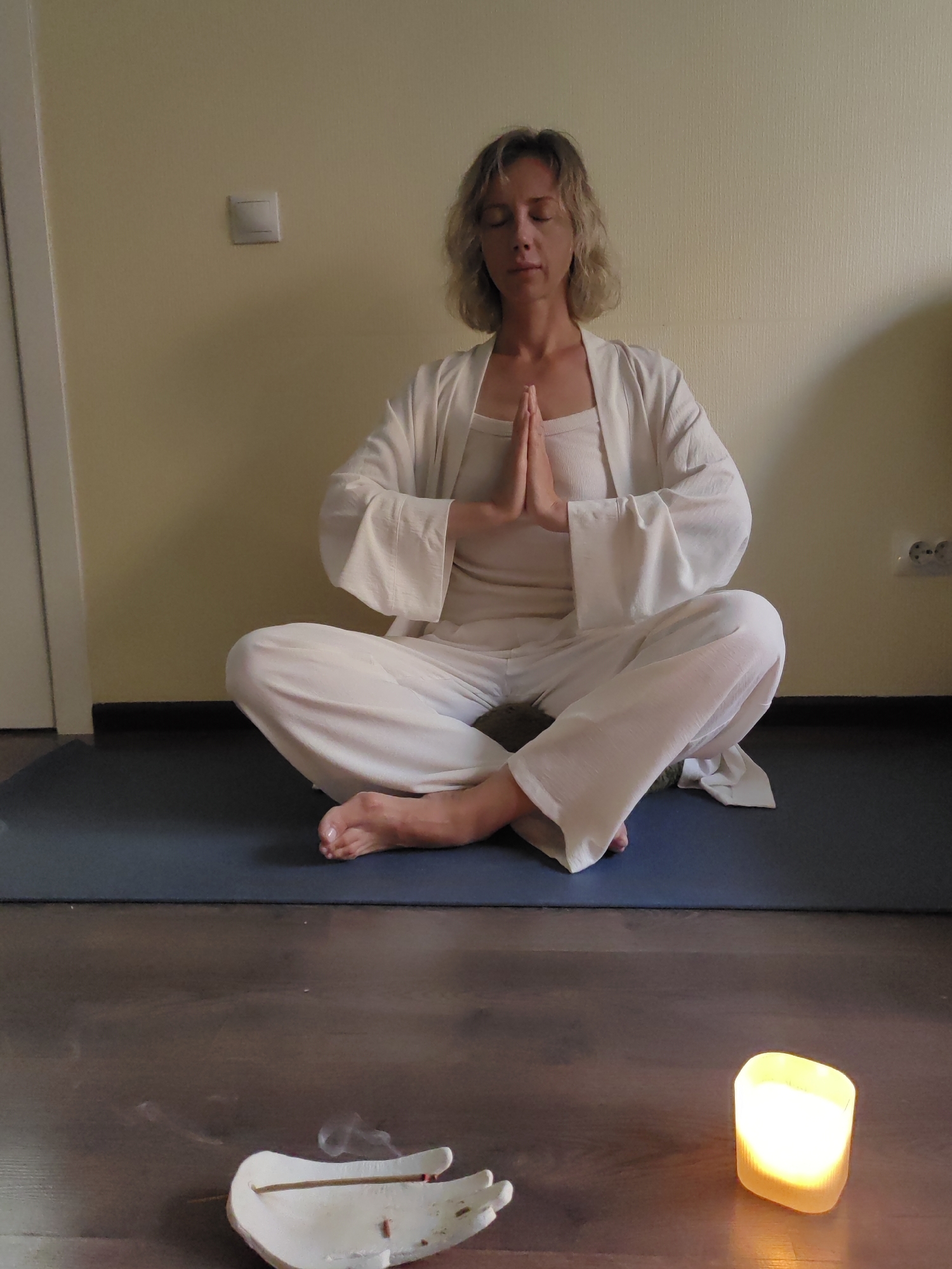 A woman with short blonde hair is meditating in a lotus position on a yoga mat. She is wearing loose-fitting white clothing, including a long-sleeved robe over a tank top and pants. Her eyes are closed, and her hands are pressed together in front of her chest. The setting appears to be an indoor space with a light yellow wall and a wooden floor. In the foreground, there is a lit candle and an incense holder with a smoldering stick, indicating a calming and spiritual atmosphere. The overall mood is peaceful and serene. The image seems to have been taken during the daytime, given the ambient light. There are no visible text elements in the image.