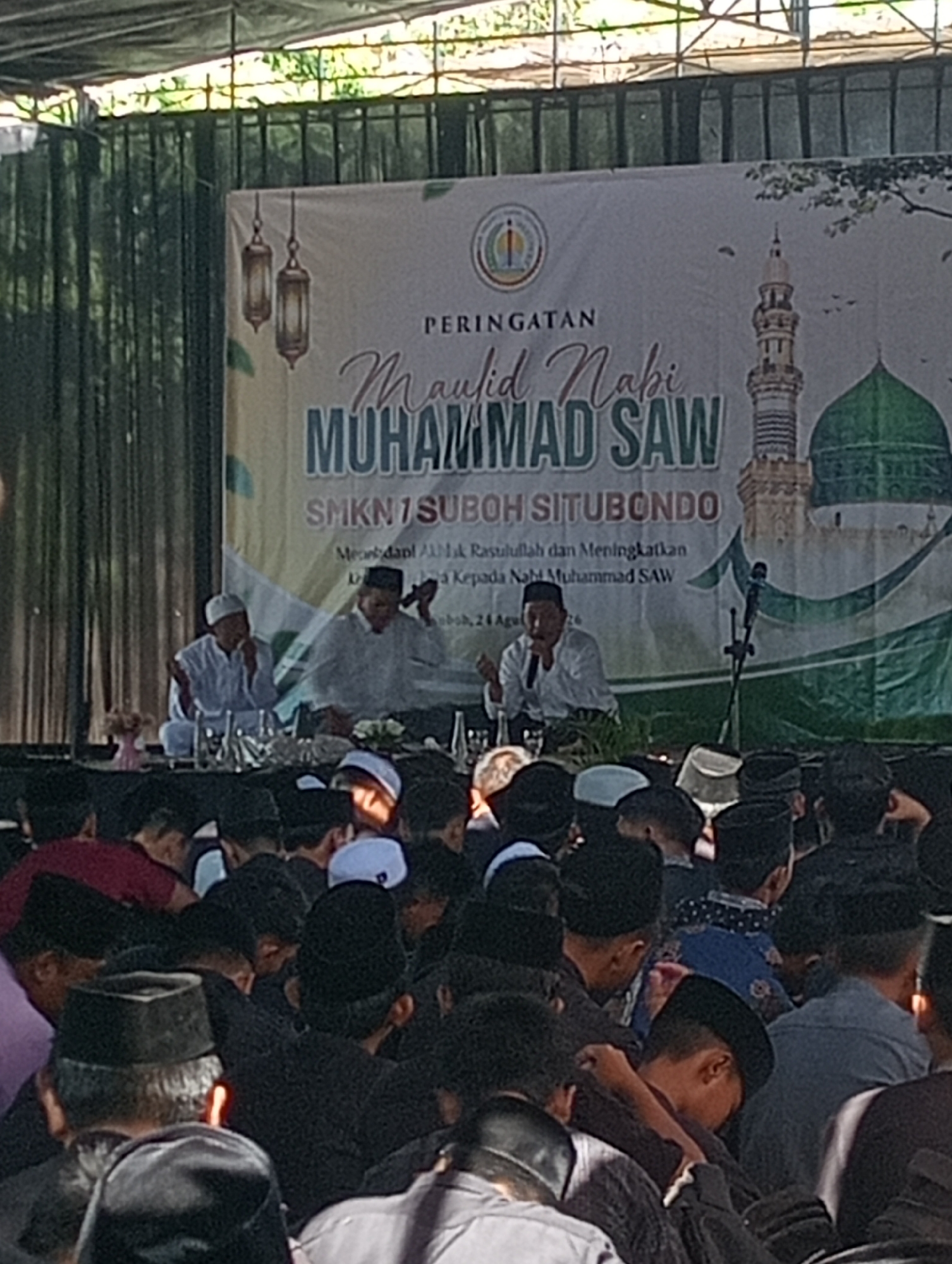 The image shows an outdoor event with a banner in the background. The banner has text that reads "PERINGATAN Maulid Nabi MUHAMMAD SAW" and "SMKN 1 SUBO SITUBONDO". There is also an illustration of a mosque and a minaret on the banner. In front of the banner, three men are seated on a stage. The man on the left is wearing a white cap and robe. The man in the middle is also wearing a white cap and robe. The man on the right is wearing a black cap and a white shirt. He is speaking into a microphone. In front of the stage, a crowd of people, mostly men, are seated, facing the stage. They are wearing various types of headwear, including peci hats. The event appears to be a religious gathering. The location is identified as Buduan, Indonesia.