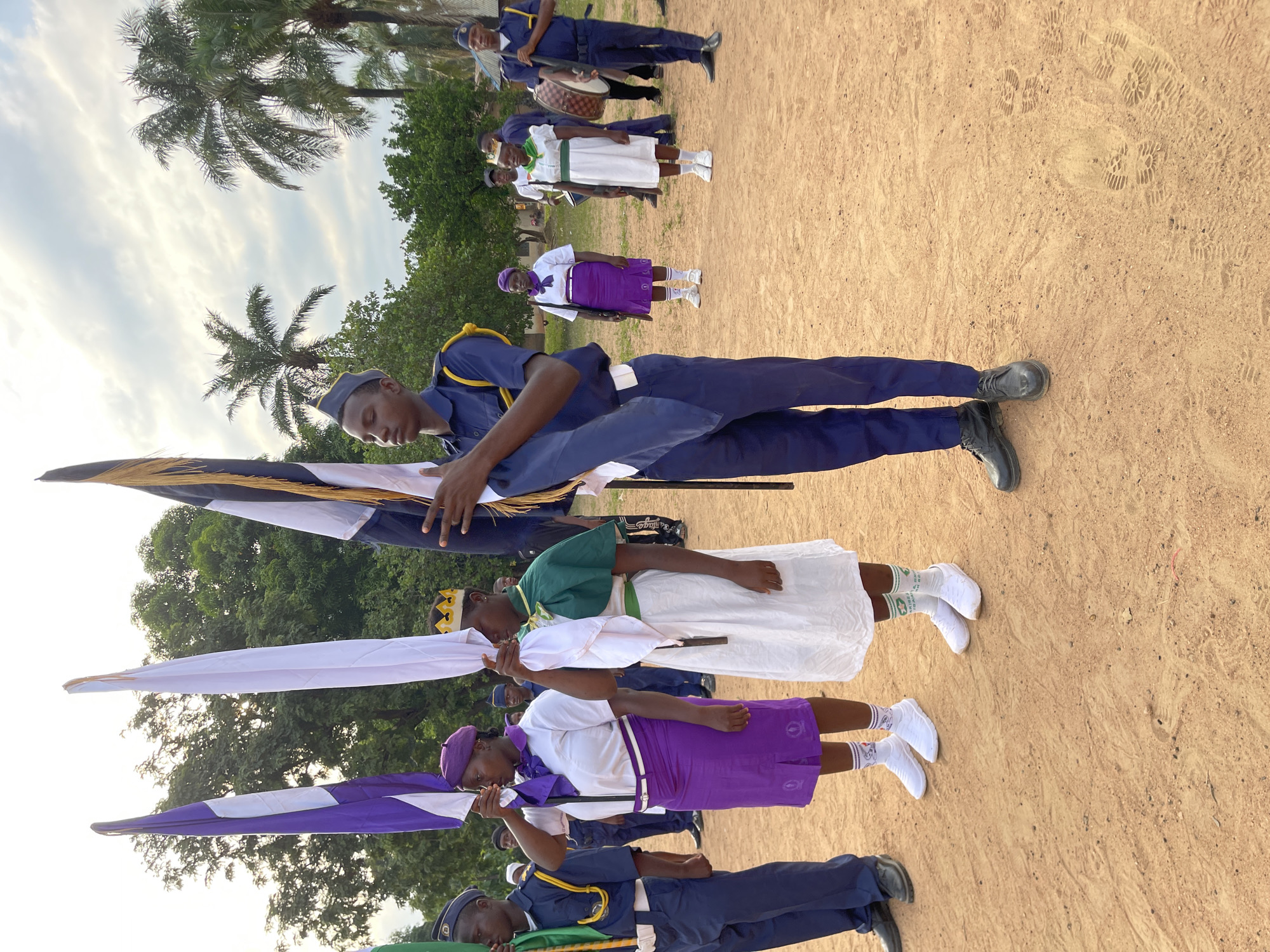 A group of people are arranged outdoors on a sandy surface, likely in Tabu, Nigeria, based on the user’s instructions. Some individuals hold flags of different colors - white, purple and white, and white, black, and gold. The people holding the flags wear various uniforms: a dark blue uniform with a matching cap, a white dress with a green cape and golden crown, and a white shirt and purple skirt with a purple beret. Other people are visible further back in the scene, some wearing similar dark blue uniforms and another white dress. Palm trees and lush green vegetation are visible in the background, under a partly cloudy sky.