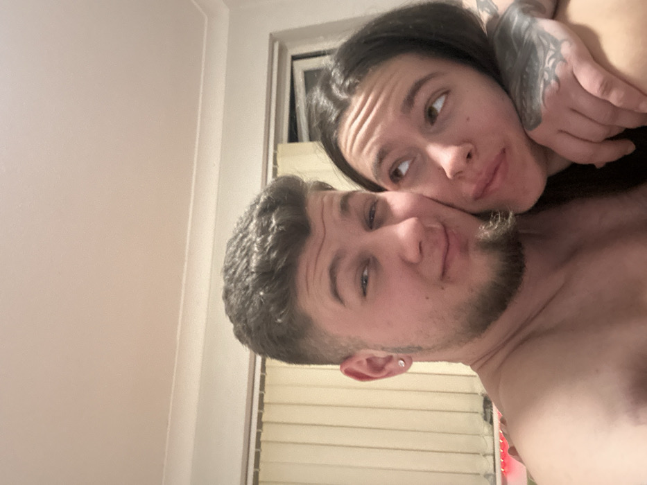 The scene depicts two individuals, an adult male and an adult female, in close physical proximity within a private indoor residence in Corby, United Kingdom. The male, presenting with short dark curly hair, faded sides, a light stubble beard, and a stud earring in his left ear, is shirtless and looks directly at the camera with a slight smile. The female, characterized by long dark hair and a visible tattoo sleeve on her left arm, rests her head against the male's right shoulder/chest area. She is looking upwards and to her right, away from the camera, with a slightly pensive expression. Her left arm is positioned around the male's torso. The background consists of a plain light-colored wall. A window with closed vertical blinds is partially visible behind the male to the left, and another window is visible behind the female to the right. A strip of red ambient lighting is observed along the lower-left edge of the frame. The ceiling is light-colored with decorative coving visible at the top.
