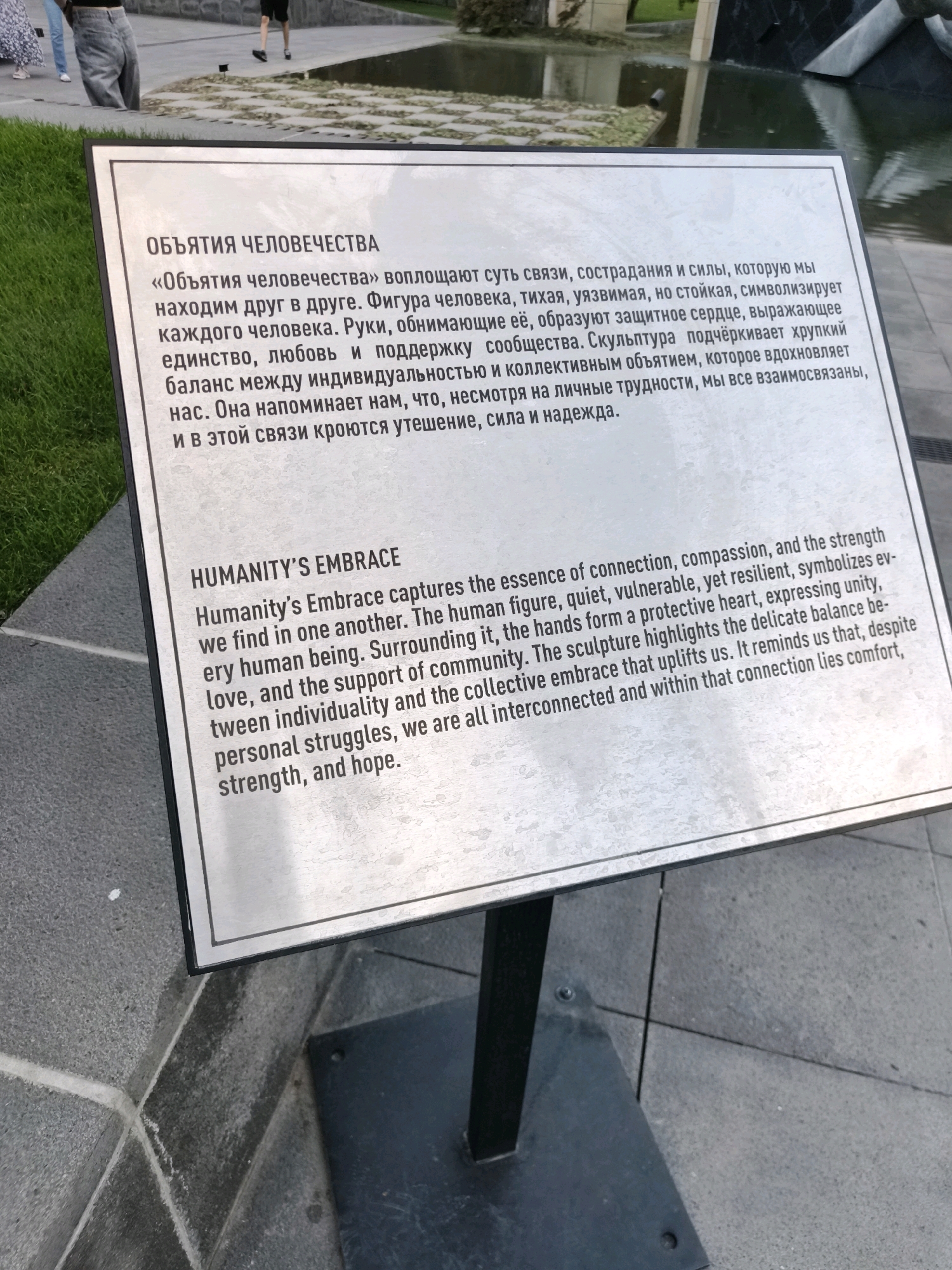 The image shows a plaque with text in Russian and English, likely an explanatory sign for a sculpture or monument. The plaque is made of brushed metal and is framed in black. The Russian text above the English text reads: "ОБЪЯТИЯ ЧЕЛОВЕЧЕСТВА" which translates to "HUMANITY'S EMBRACE". The English text below it states: "HUMANITY'S EMBRACE Humanity's Embrace captures the essence of connection, compassion, and the strength we find in one another. The human figure, quiet, vulnerable, yet resilient, symbolizes ev-ery human being. Surrounding it, the hands form a protective heart, expressing unity, love, and the support of community. The sculpture highlights the delicate balance be-tween individuality and the collective embrace that uplifts us. It reminds us that, despite personal struggles, we are all interconnected and within that connection lies comfort, strength, and hope." The plaque is positioned outdoors, on a gray, textured surface, possibly a paved area in a park or public square. In the background, parts of a water feature and people are visible. The overall scene suggests an urban public space dedicated to art or commemoration.