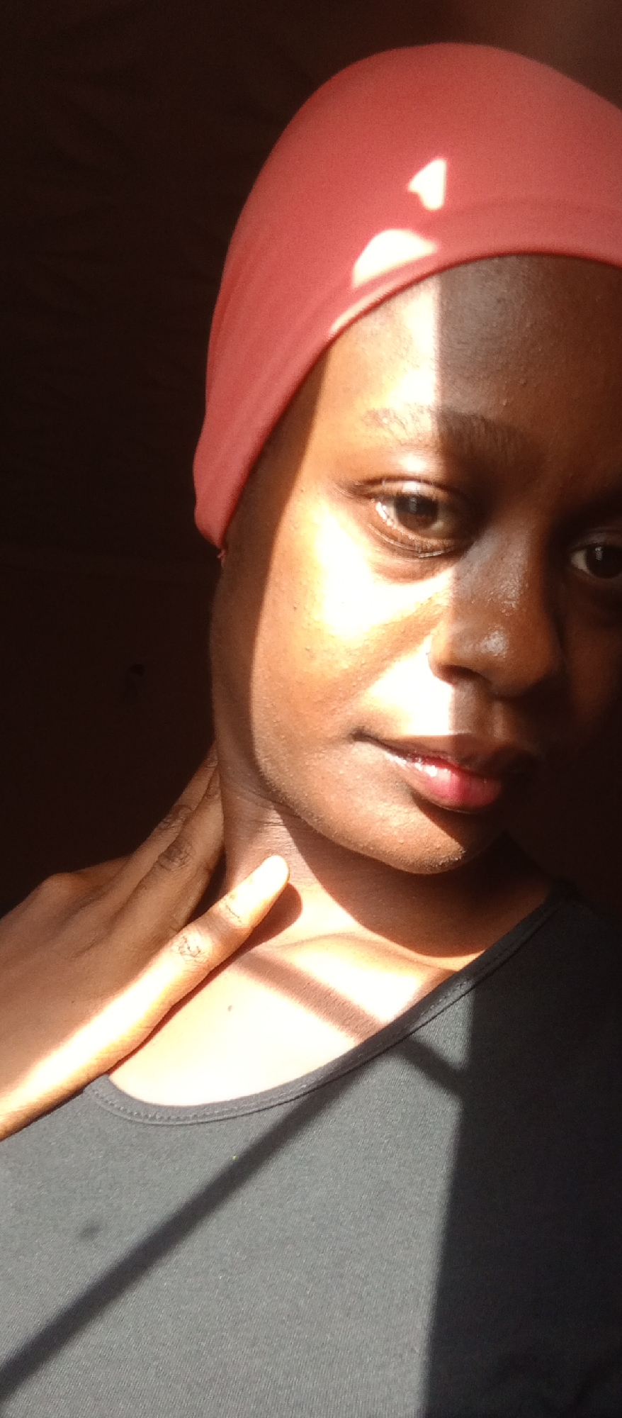 A close-up portrait shows a Black woman with short dark hair covered by a light red headscarf. Her dark gray t-shirt is partially obscured by shadow. She is touching her neck with her right hand. The subject's face is illuminated by a strong light source, casting harsh shadows and highlights on her skin. The background is mostly dark. There is no information provided in the image to suggest that the scene is in Akwanga, Nigeria. The photograph is not forensic.