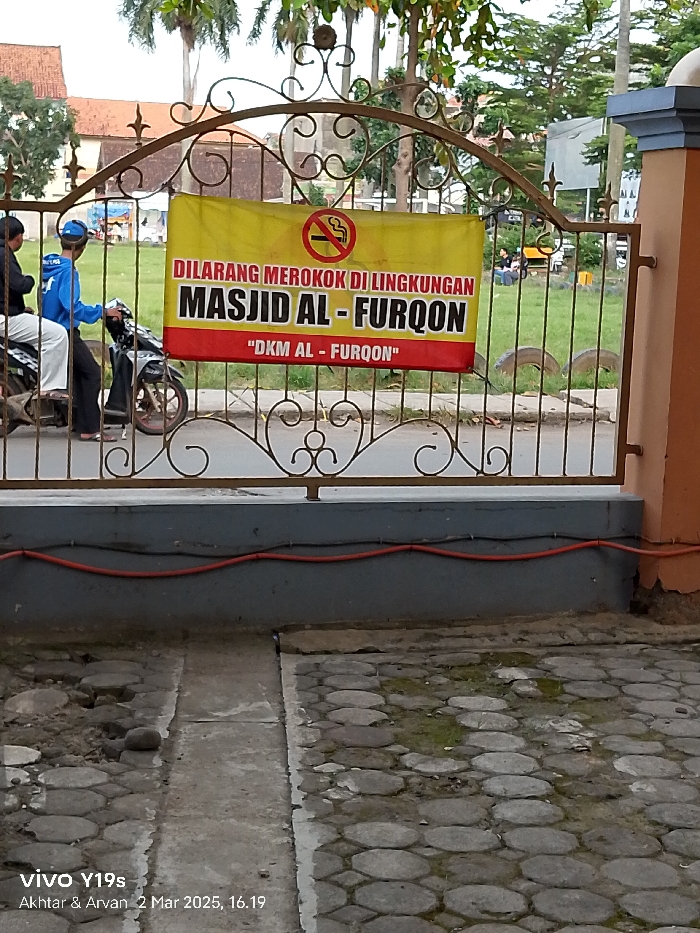A sign in Indonesian reads "DILARANG MEROKOK DI LINGKUNGAN MASJID AL - FURQON", which translates to "Smoking is prohibited in the area of MASJID AL - FURQON", located in Kecamatan Haurgeulis, Indonesia (latitude: -6.46, longitude: 107.943). The sign is mounted on a black metal gate with a decorative scroll design. A red and white "no smoking" symbol is visible on the sign. Two people are riding a motorcycle in the background. The motorcycle is blue with a black seat and a red frame. The pavement in the foreground is made of large, irregularly shaped gray cobblestones. There is a red electrical wire running along the base of a concrete wall in the foreground. The scene is set in an outdoor area with green grass and trees in the background.
